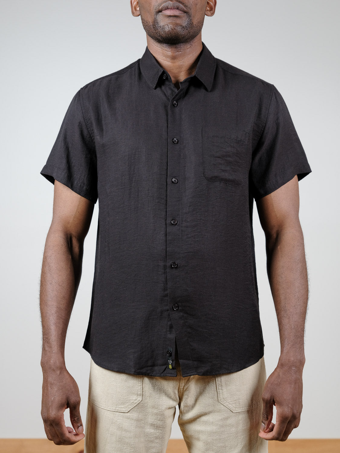 A person wearing the Hiker Short Sleeve Shirt by ID Menswear in black linen with a chest pocket and beige pants, standing against a plain light background. The face is not fully visible—ideal for effortless summer basics.