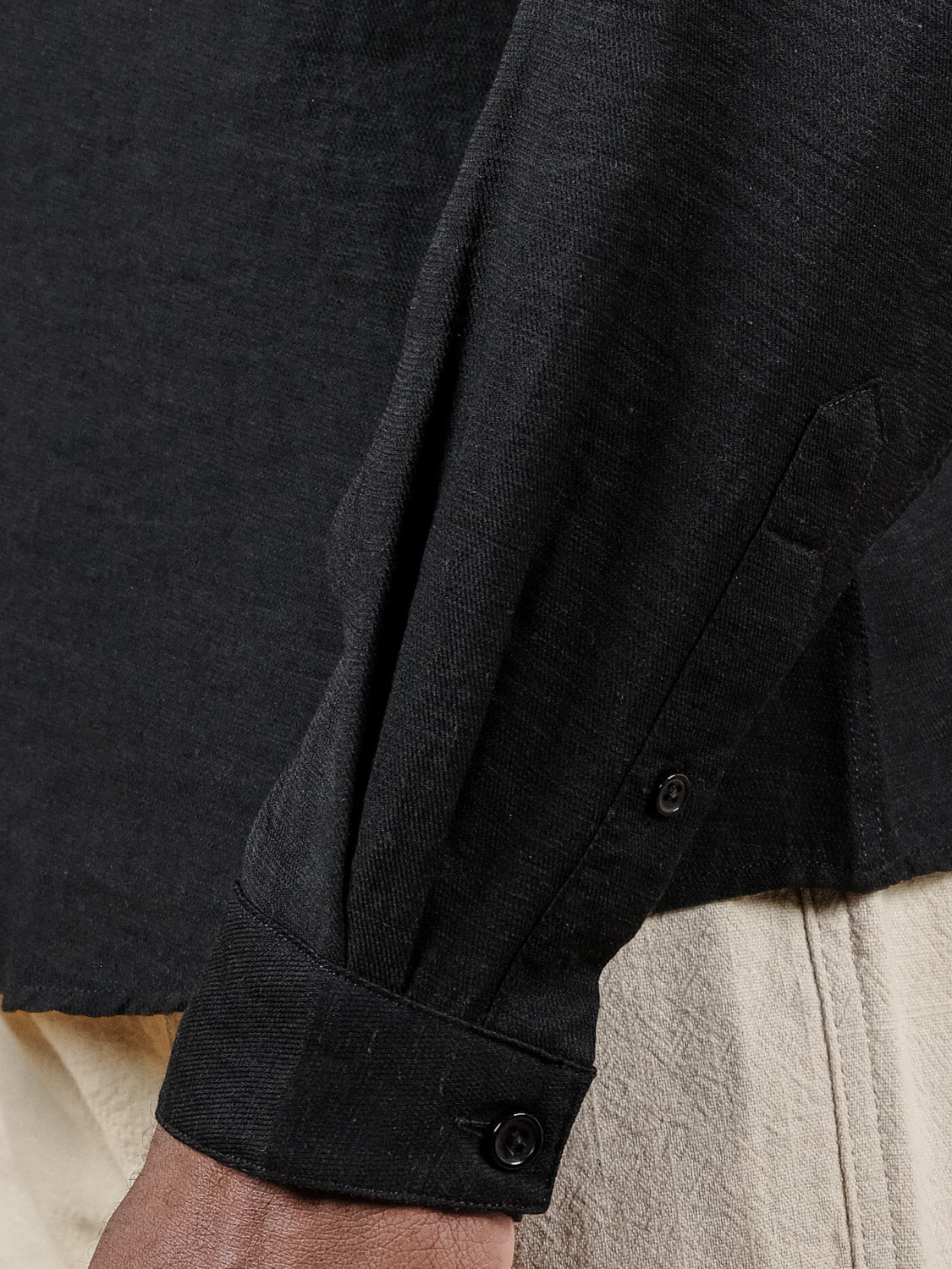 Close-up of a person wearing the ID Menswear Lagos Long Sleeve Shirt in black with buttoned cuffs, paired with beige pants. The shirt’s relaxed fit and details are shown, with the person’s hand partially visible at the image’s bottom.