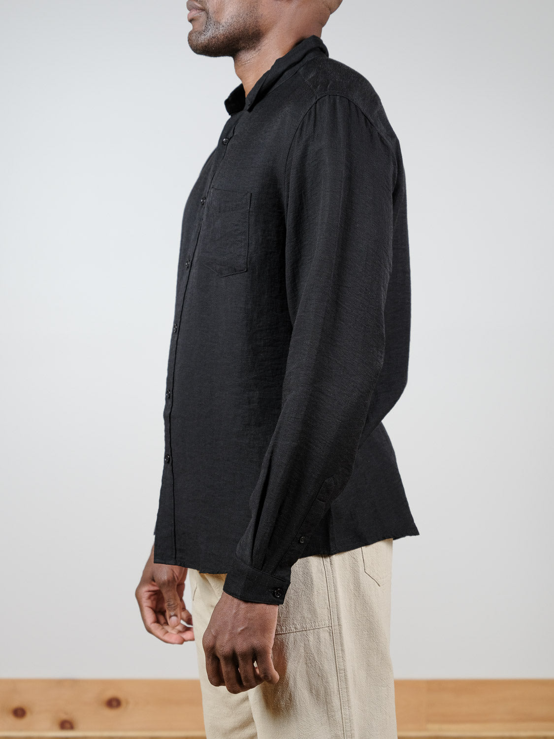 A person wears an ID Menswear Lagos Long Sleeve Shirt in black with light beige pants, standing sideways against a plain light background, their left hand seen and relaxed at their side.