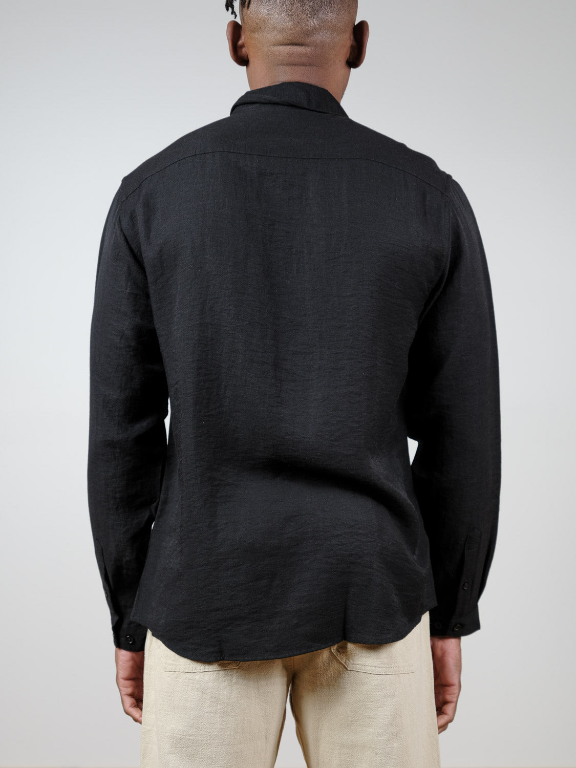 A person is seen from behind, wearing a relaxed fit black Lagos Long Sleeve Shirt by ID Menswear and light beige pants, standing against a plain light background.