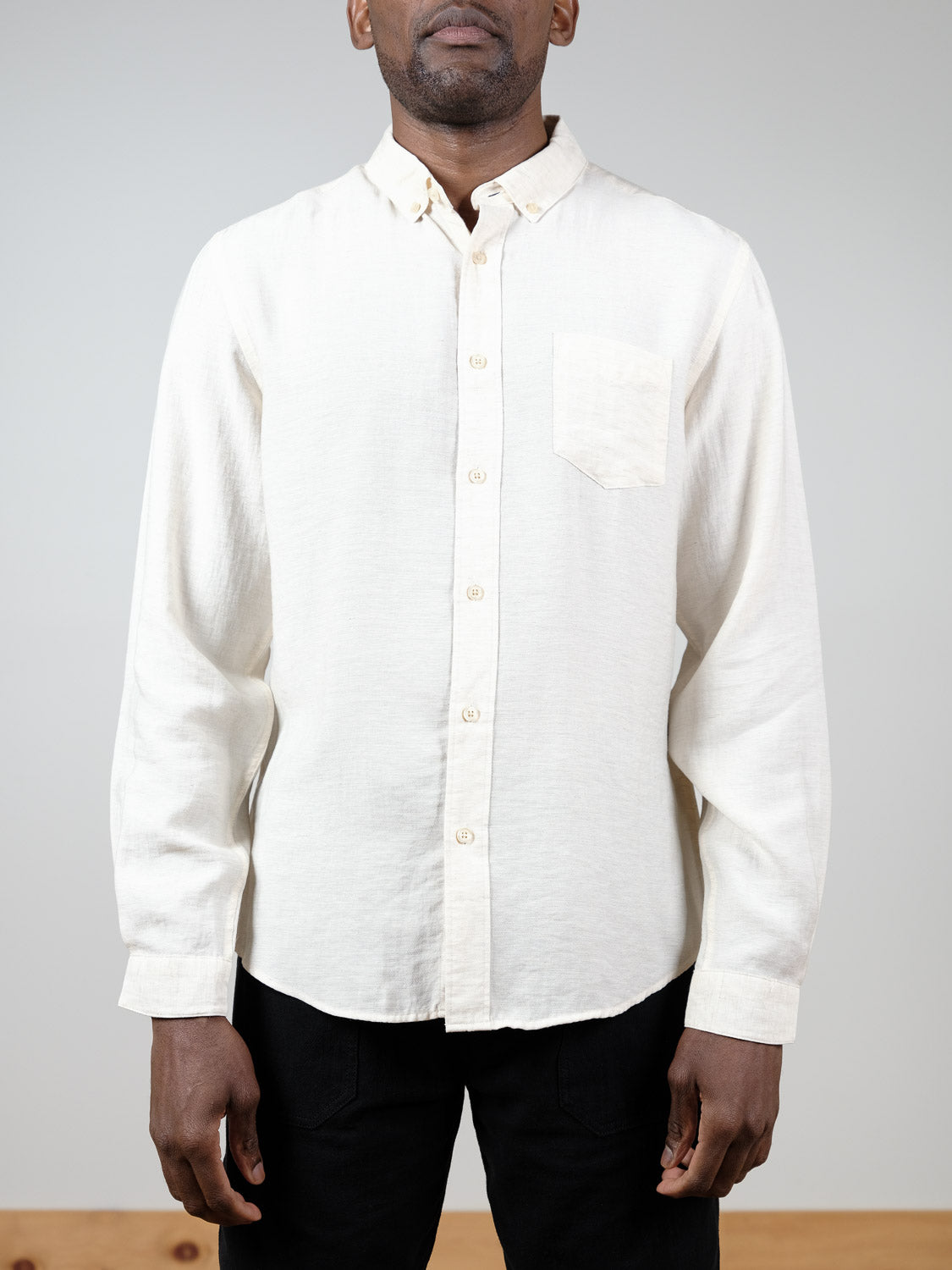 A person in a relaxed fit, cream Lagos Long Sleeve Shirt by ID Menswear with a chest pocket stands against a plain light background, only their upper body visible.