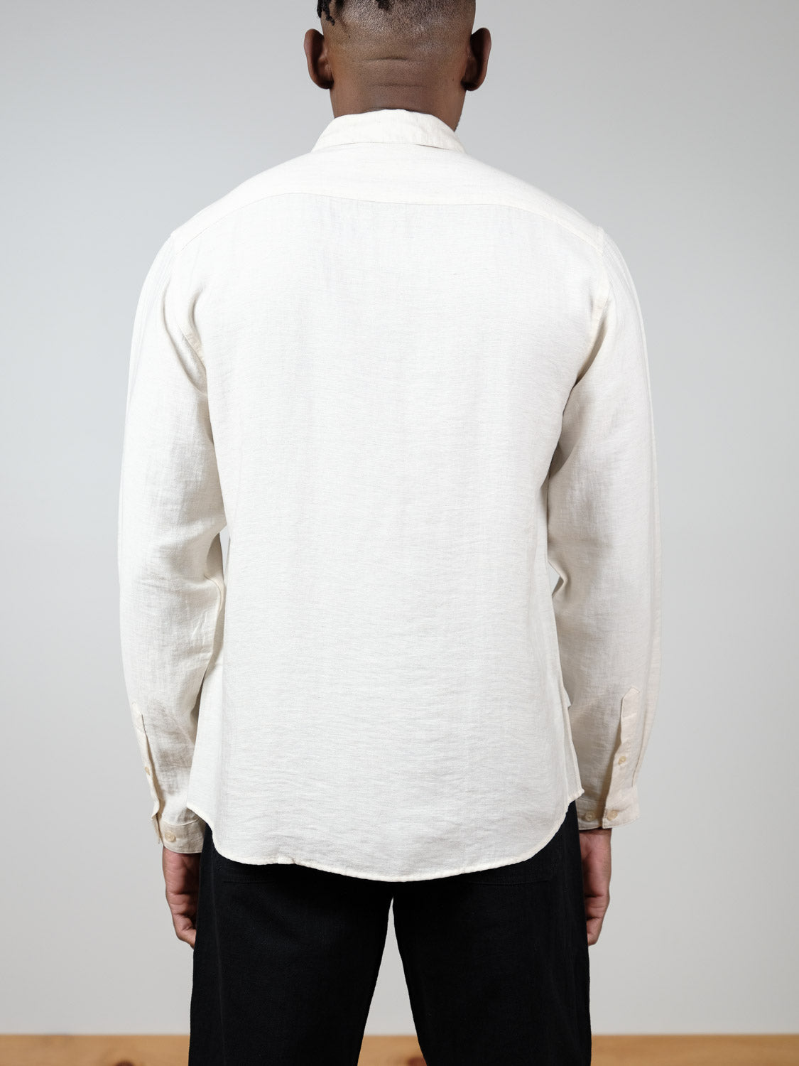 A person faces away from the camera, wearing a relaxed fit cream ID Menswear Lagos Long Sleeve Shirt and black pants against a plain light gray background.