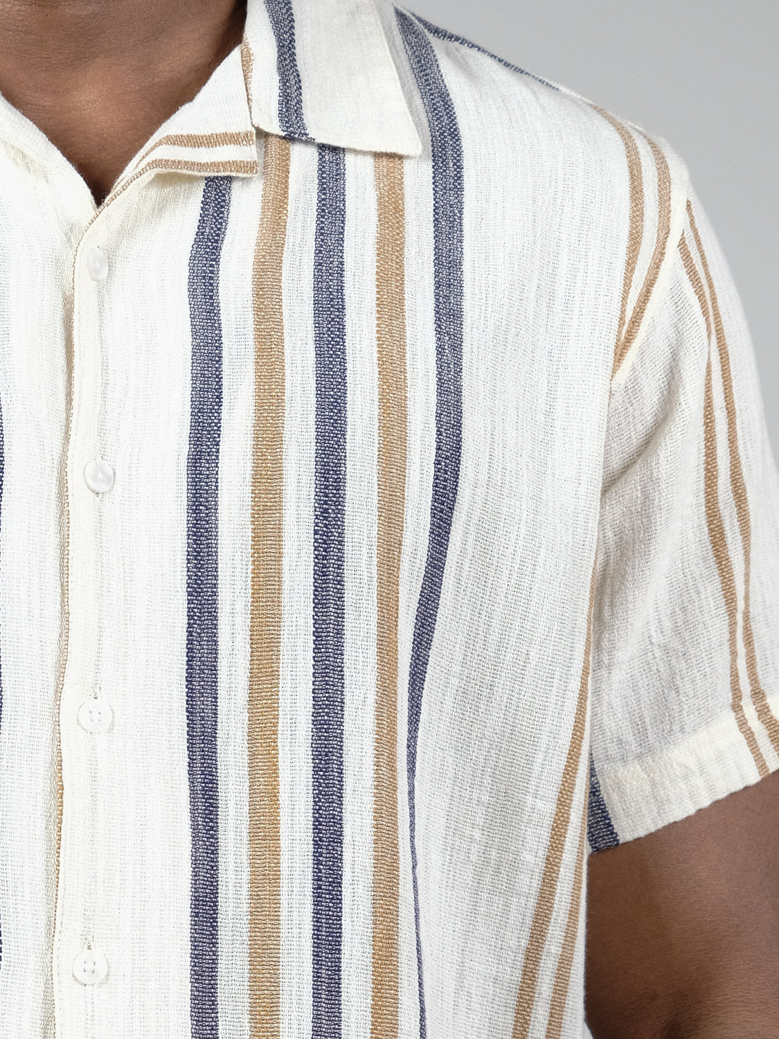 A close-up of a person wearing the !Solid Isai Short Sleeve Shirt, featuring vertical blue, tan, and white stripes, a camp collar, cotton fabric, and buttoned front. Only part of the upper body is visible.