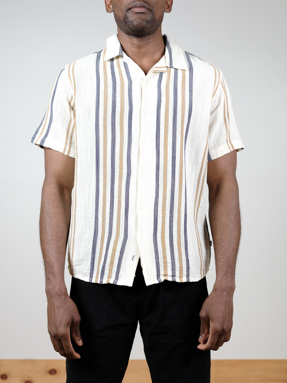 A man stands against a plain background wearing the !Solid Isai Short Sleeve Shirt, featuring vertical beige, blue, and white stripes with a camp collar. He pairs the cotton shirt with black pants; his face is partially out of frame.