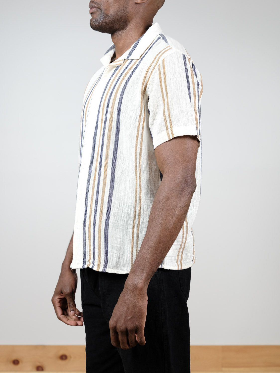A man stands in profile against a plain background, wearing the !Solid Isai Short Sleeve Shirt with vertical beige, blue, and brown stripes, paired with black pants.