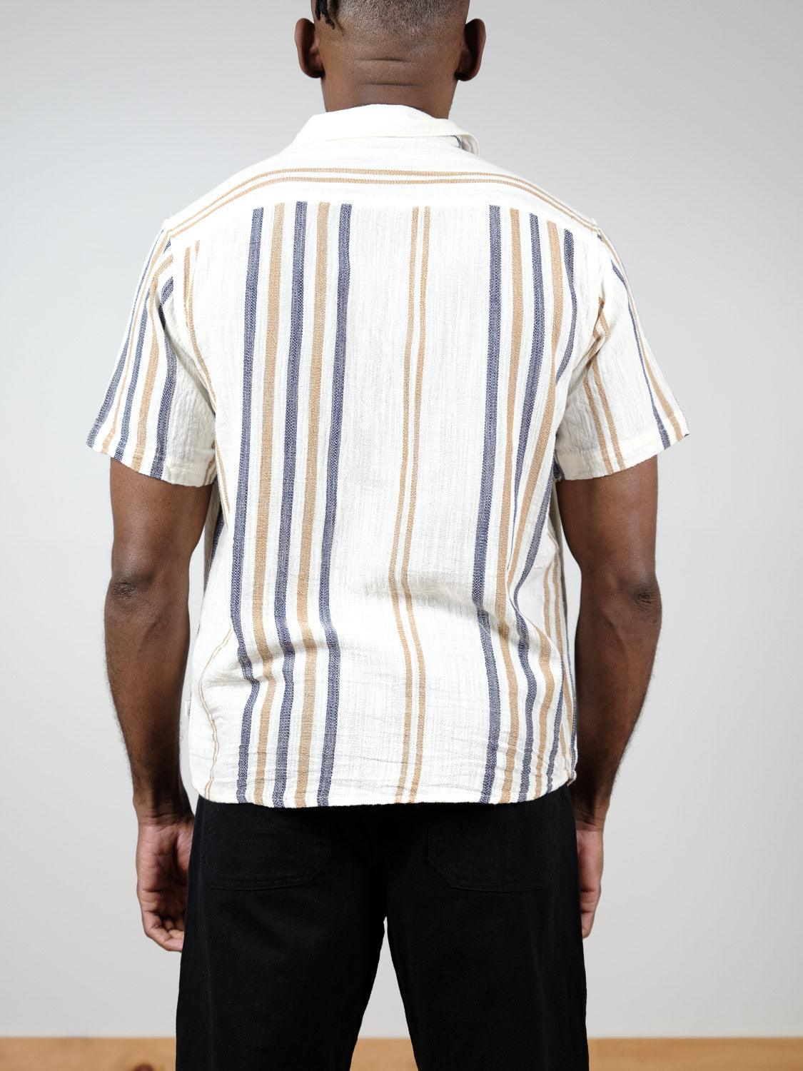 A person faces away from the camera, wearing the Isai Short Sleeve Shirt by !Solid—a cream cotton striped camp-collar shirt—paired with black pants against a clean, neutral backdrop.