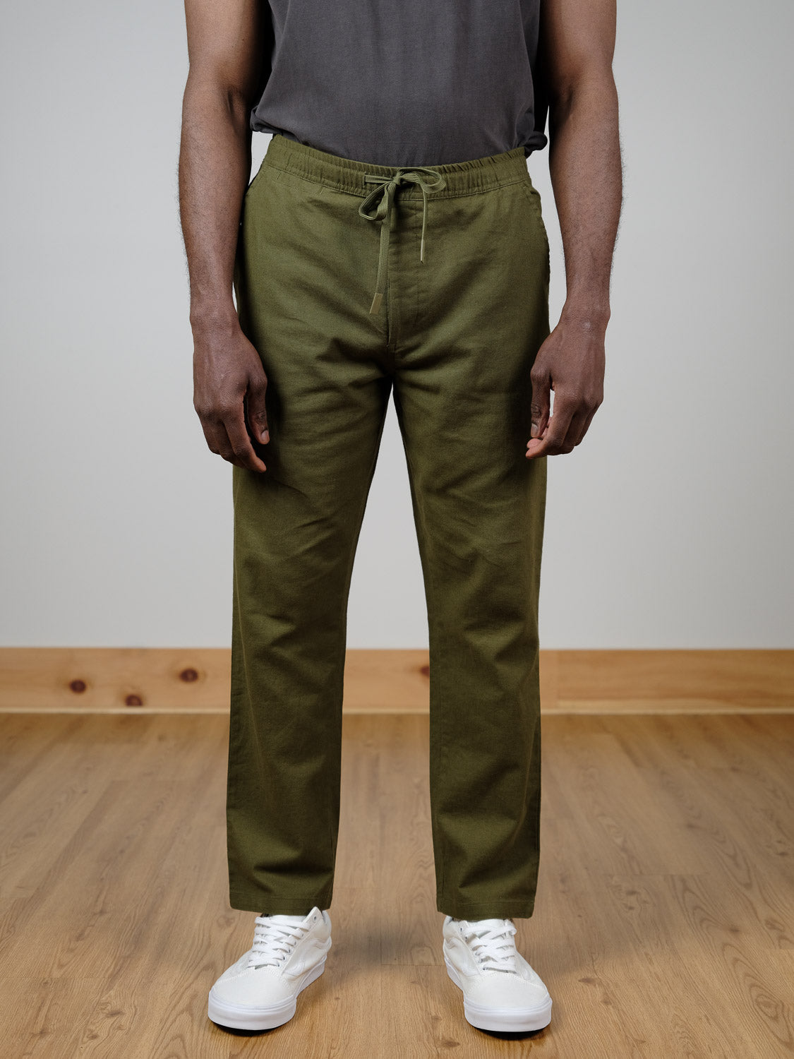 A person wears the Laguna Linen Cotton Drawstring Pant by ID Menswear in a relaxed crop fit, paired with a dark gray t-shirt and white sneakers, standing on a light wood floor against a plain light-colored wall.