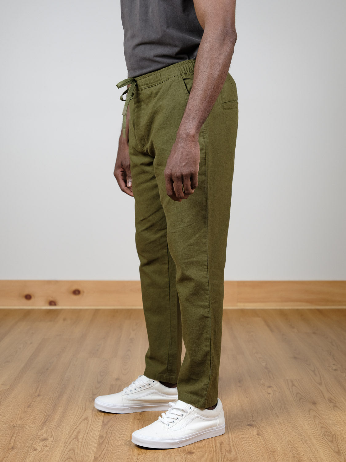 A person stands indoors on a wooden floor, wearing olive green Laguna Linen Cotton Drawstring Pants by ID Menswear, a dark gray t-shirt, and white sneakers. The side view image shows them from the shoulders down.