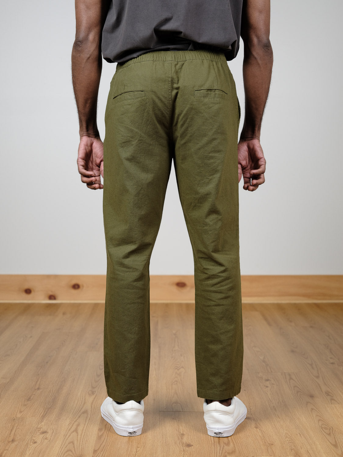 A person stands indoors on a wooden floor, shown from the back, wearing ID Menswear’s olive green Laguna Linen Cotton Drawstring Pant and white sneakers. Their arms hang by their sides, and a dark shirt is partially visible.