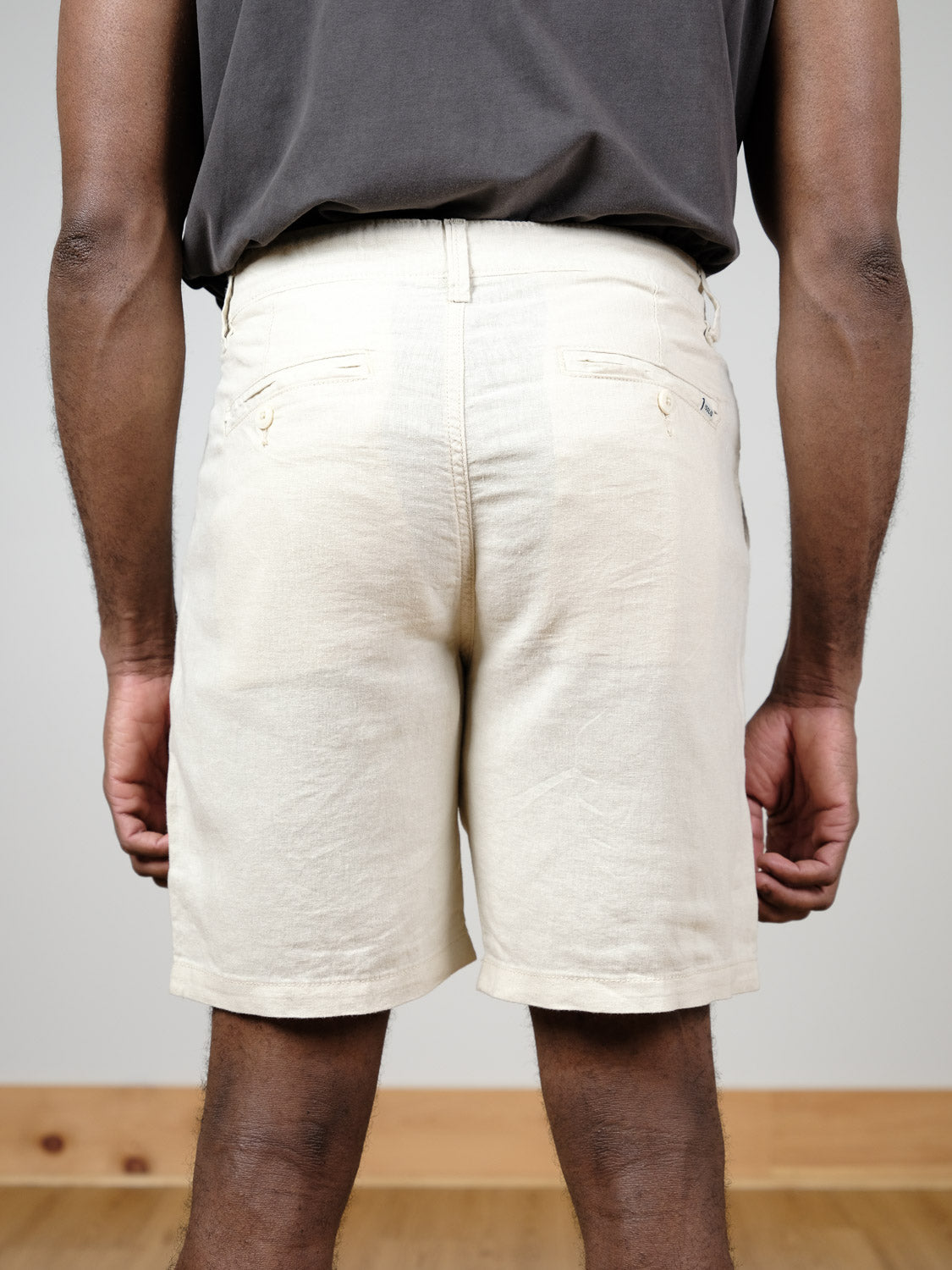 A person is shown from the waist down, wearing the !Solid Aurelius Short in light beige linen with back pockets and a dark shirt, standing indoors on a wooden floor against a neutral wall.