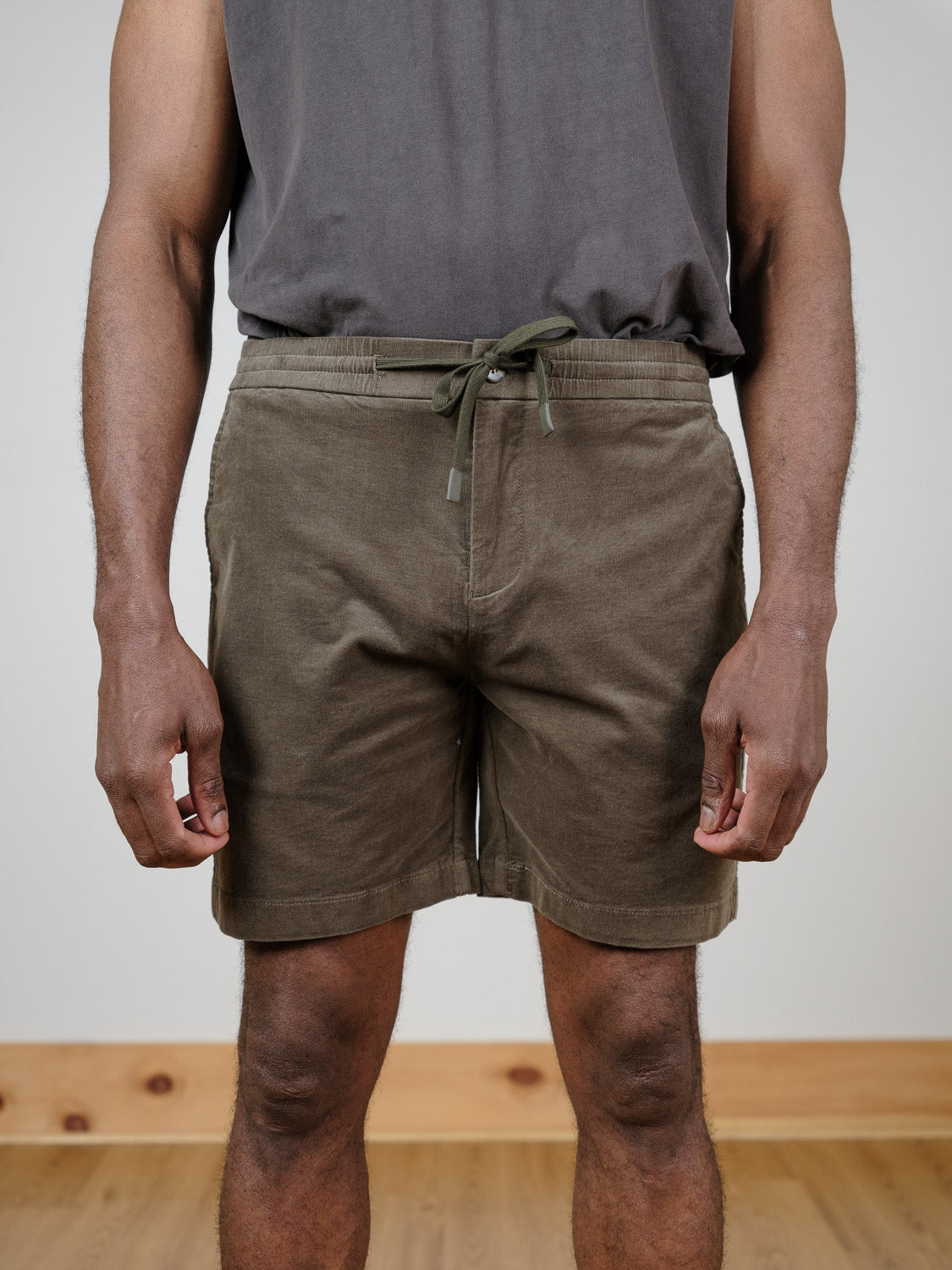 A person stands indoors on a wooden floor, arms relaxed at their sides, wearing a dark sleeveless shirt and olive green Odyssey Corduroy Stretch Shorts by ID Menswear with an elastic waistband.
