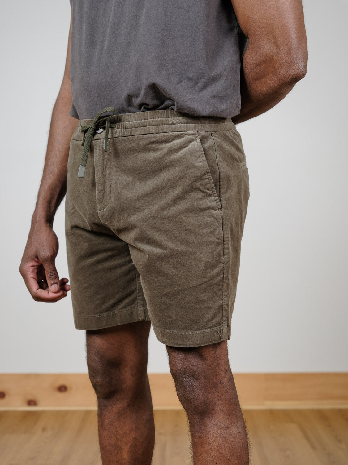 A person stands indoors on a wooden floor, hands in pockets, wearing an untucked dark gray t-shirt and Odyssey Corduroy Stretch Short by ID Menswear in olive green, shown from shoulders to knees.