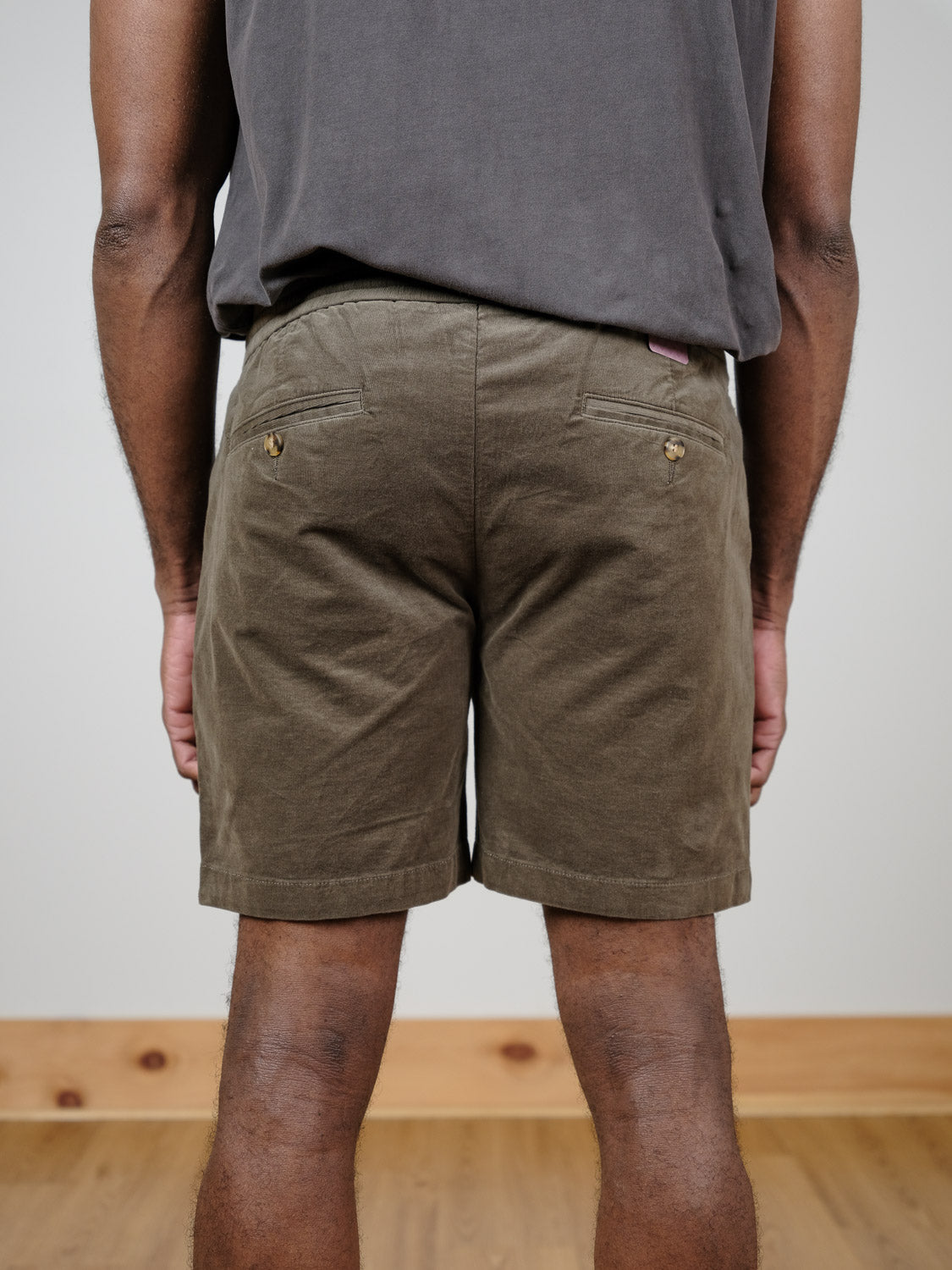 A person faces away from the camera indoors, wearing ID Menswear’s Odyssey Corduroy Stretch Short in olive green with an elastic waistband and a dark shirt. The wooden floor contrasts with the plain light-colored wall.