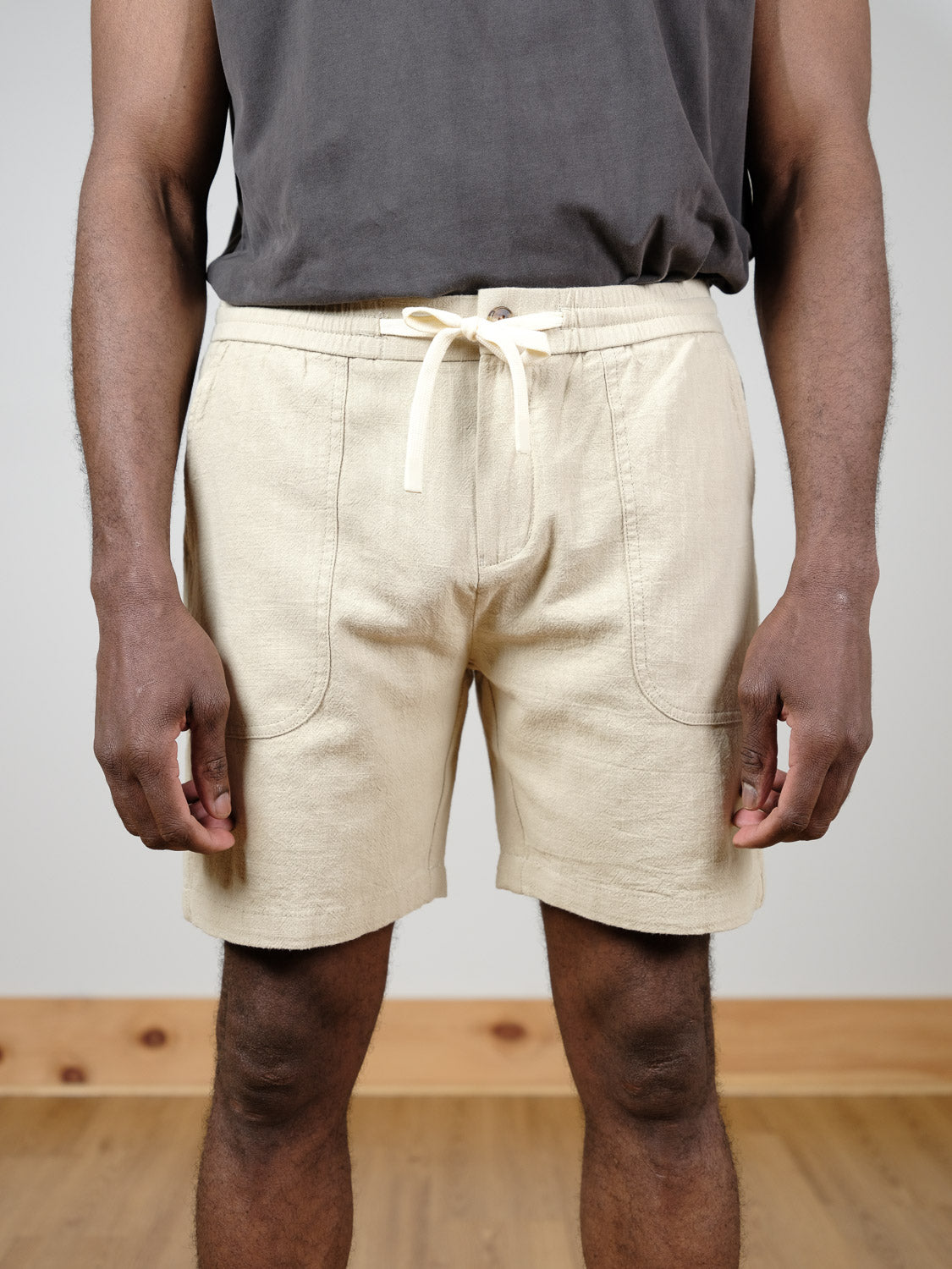 A person stands indoors on a wooden floor, arms relaxed, wearing a dark gray t-shirt and ID Menswear's Phi Phi Linen Blend Drawstring Short in light beige. The background is plain with a wooden baseboard.