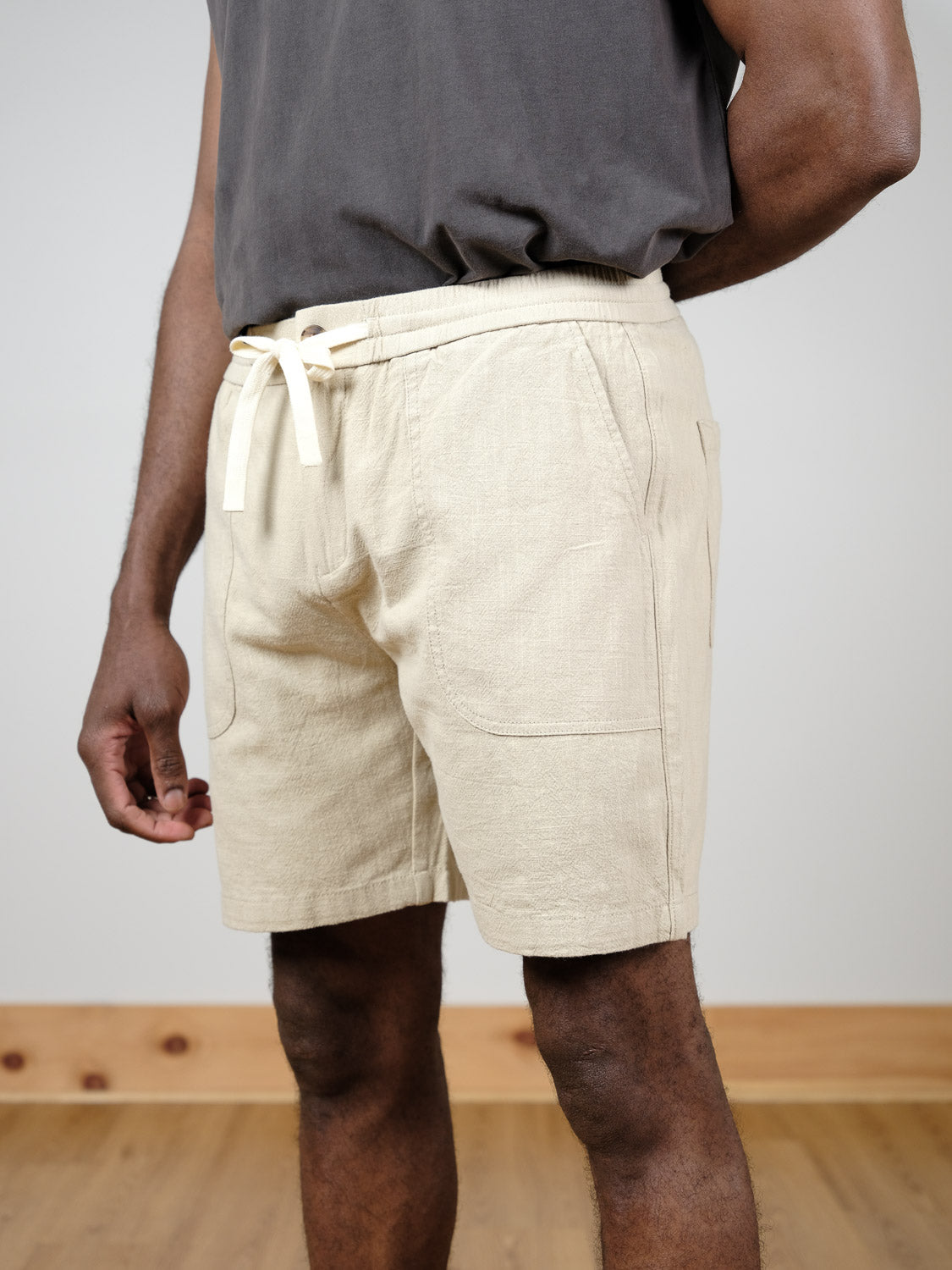 A person in a dark gray shirt and ID Menswear's Phi Phi Linen Blend Drawstring Short in light beige stands indoors on a wooden floor. The relaxed-fit shorts feature an elastic waistband and side pockets for comfort and convenience.