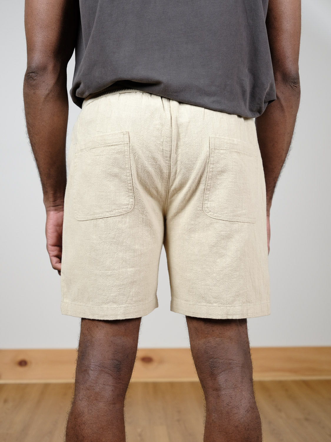 A person stands indoors on a wooden floor, shown from the waist down and from behind, wearing ID Menswear's Phi Phi Linen Blend Drawstring Short paired with a dark gray shirt. The shorts feature an elastic waistband and two back pockets.