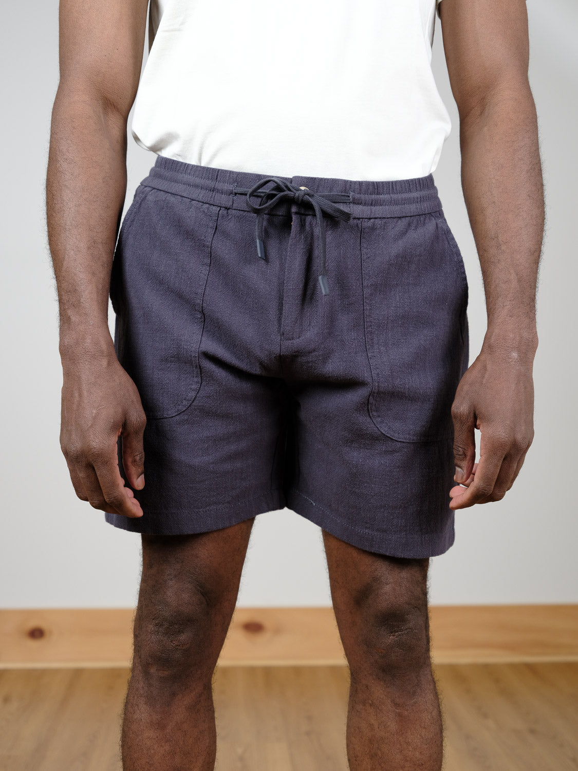 A person stands indoors on a wooden floor wearing a white shirt and ID Menswear’s Phi Phi Linen Blend Drawstring Short in dark blue. The image is cropped to show only their torso and legs, with hands relaxed at their sides.