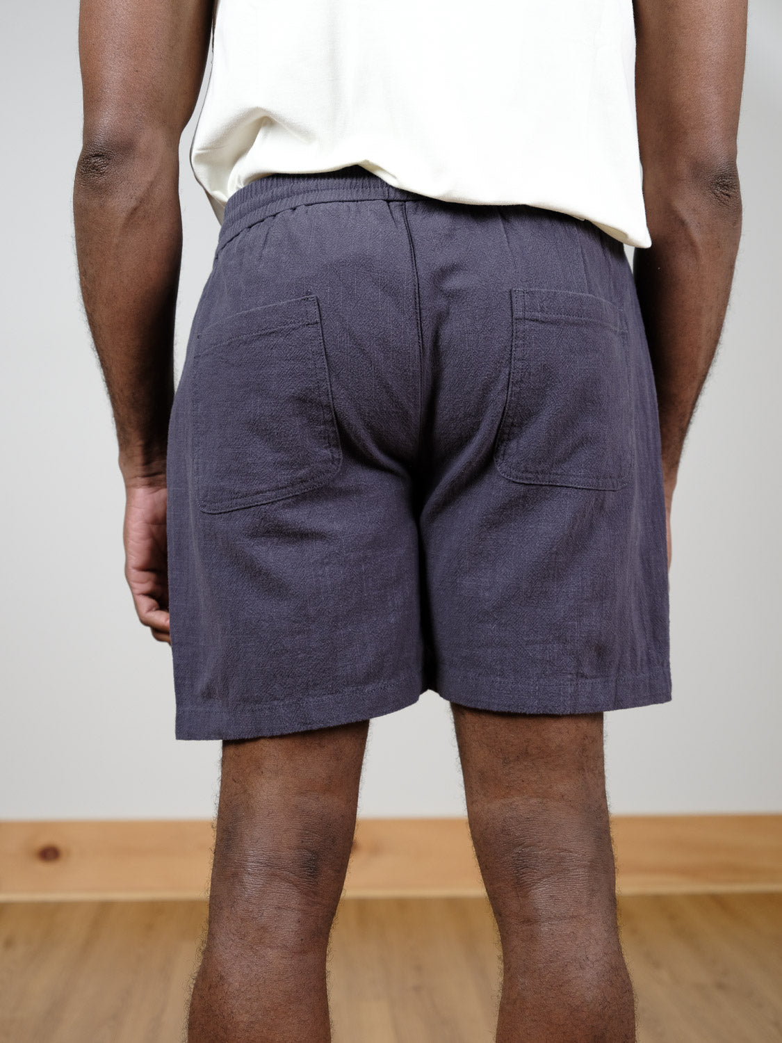 A person stands indoors with their back to the camera, wearing a white shirt and ID Menswear's Phi Phi Linen Blend Drawstring Short in dark gray, featuring an elastic waistband, two back pockets, and slightly above knee length.