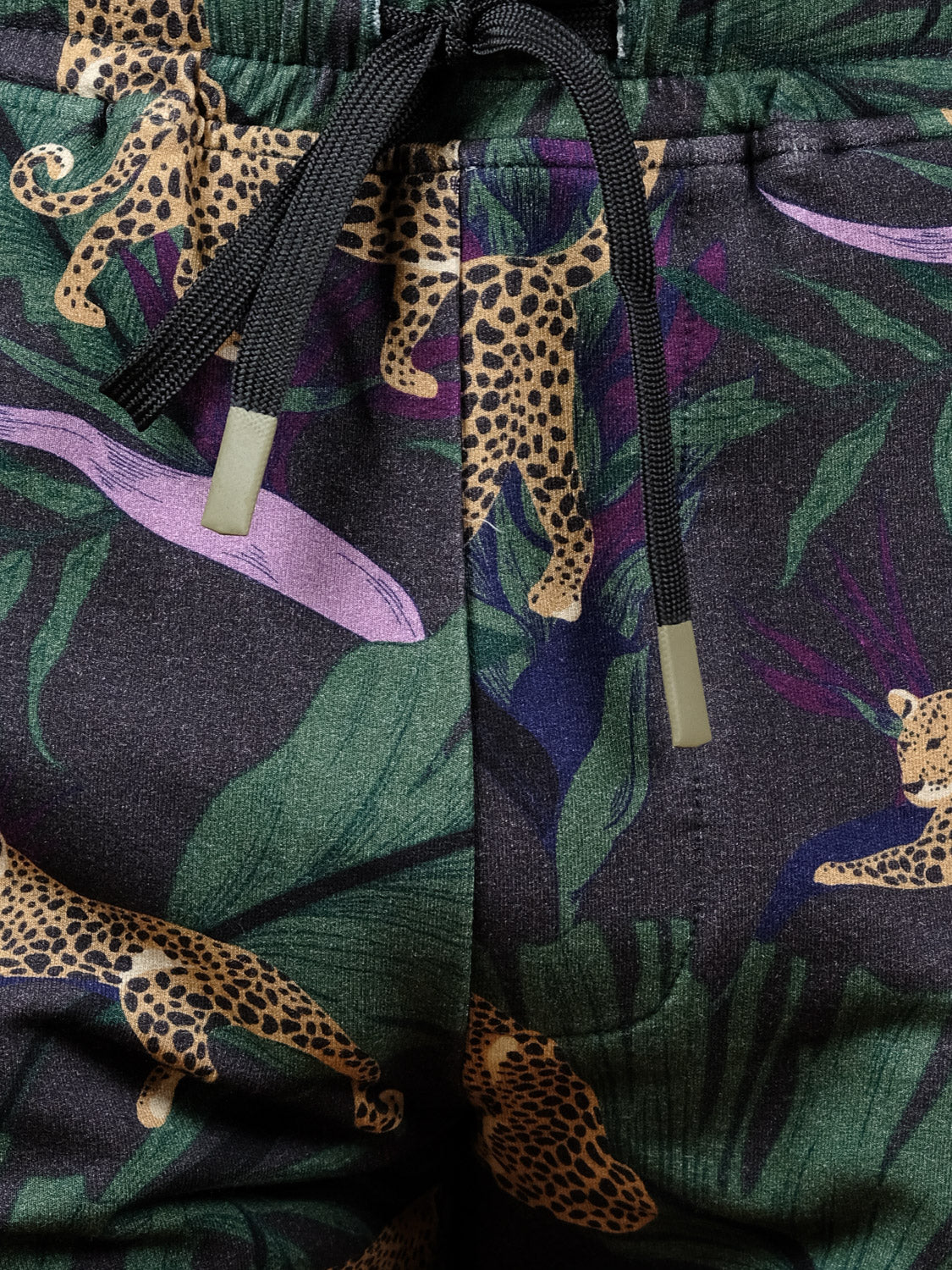 Close-up of the Samui Leopard Print Short by ID Menswear, made from organic cotton with a vibrant jungle leopard pattern, green leaves, and purple accents. Black drawstrings with metal tips are visible at the waistband.