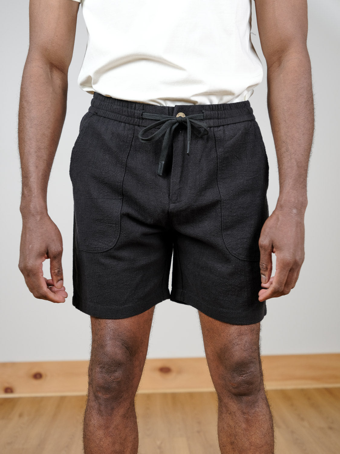 A person stands indoors on a wooden floor wearing a white T-shirt and ID Menswear's Phi Phi Linen Blend Drawstring Short, hands relaxed at their sides.