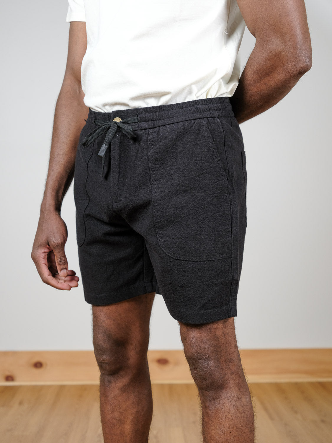 A person wears a white t-shirt and black Phi Phi Linen Blend Drawstring Shorts from ID Menswear, standing indoors on a wooden floor with their left hand on their hip and right arm relaxed by their side.