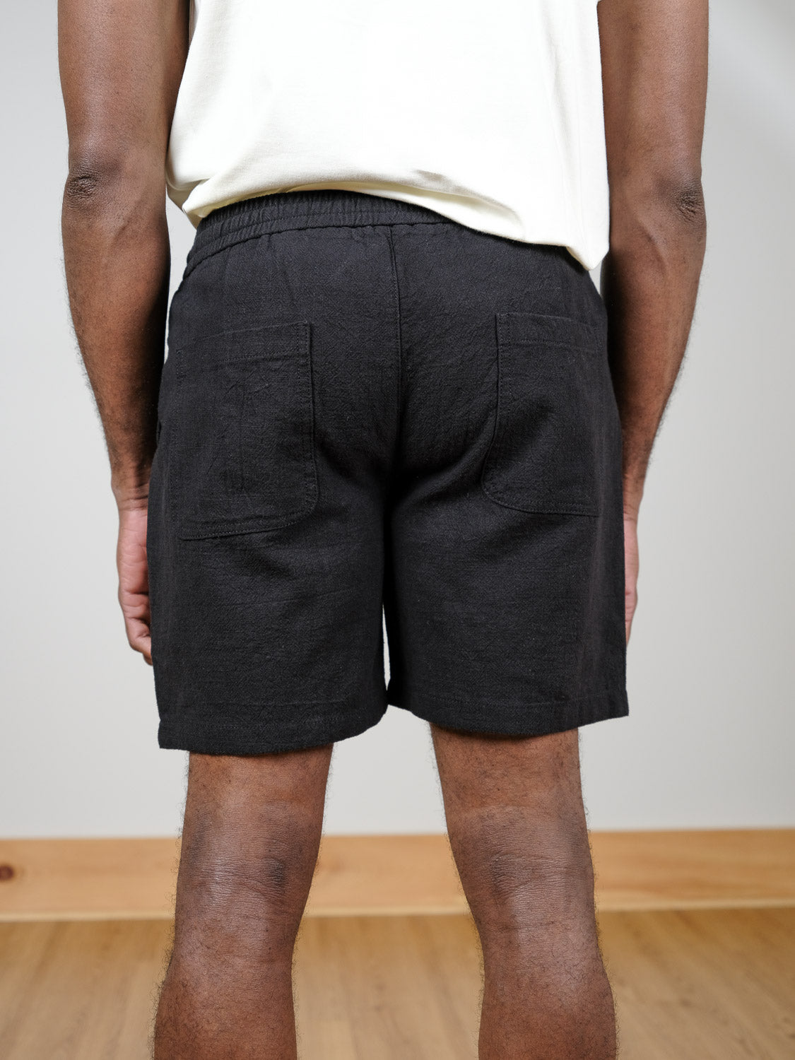 A person stands indoors facing away from the camera, wearing a cream shirt and black Phi Phi Linen Blend Drawstring Shorts by ID Menswear, which feature two back pockets. The background shows a light wall and wooden floor.