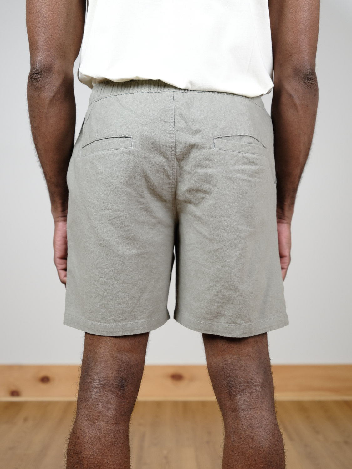 Someone wearing the Kotao Linen Blend Drawstring Short by ID Menswear in light gray and a white shirt stands indoors with their back to the camera, on a wooden floor against a plain light-colored wall.