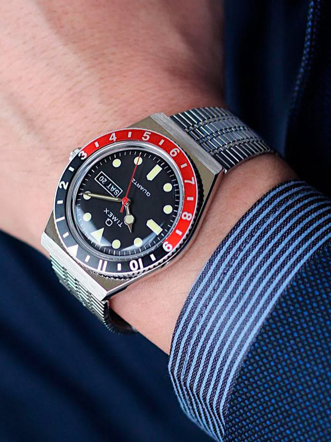 A close-up of a wrist wearing the Timex Q Timex Reissue 38mm Stainless Steel Bracelet Watch featuring a black dial, red and black bezel, day-date display, and paired with a dark striped shirt and blue jacket.