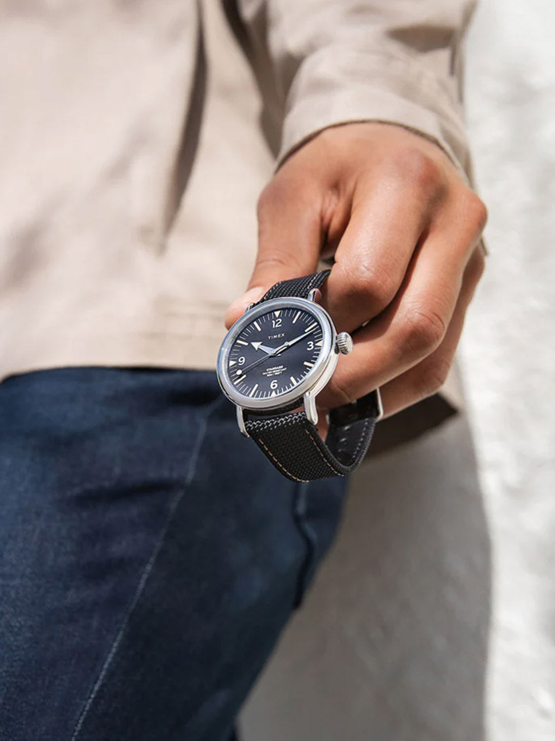 A person in a beige jacket and blue jeans holds a Timex Standard 40mm Fabric Strap Watch. The military-inspired piece features an interchangeable black strap and dark face set to 10:10, against a blurred white wall backdrop.