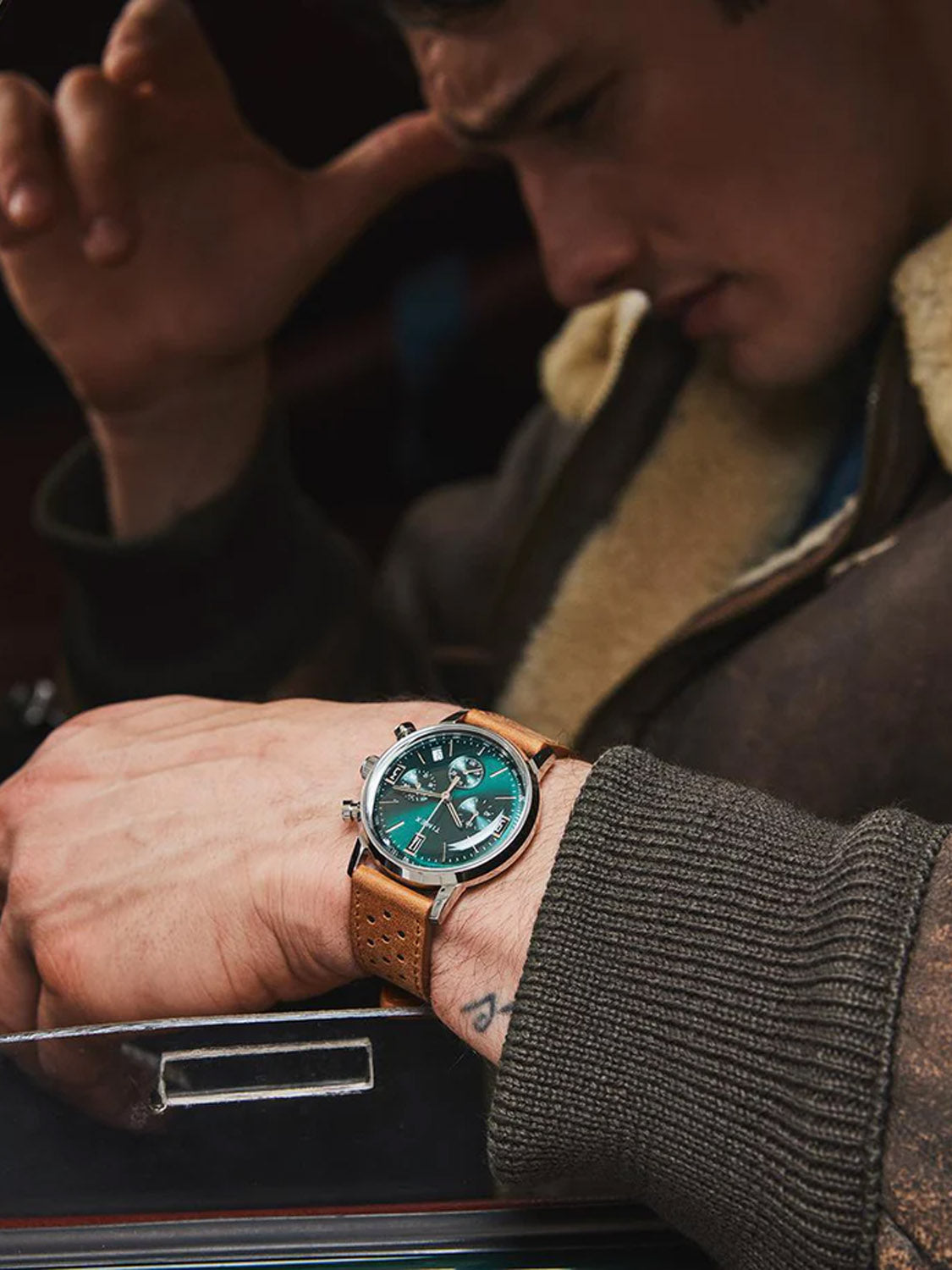 A person in a brown jacket, looking down thoughtfully with their arm on a metal surface, wears a Timex Marlin® Chronograph Tachymeter featuring a green dial and brown leather strap.