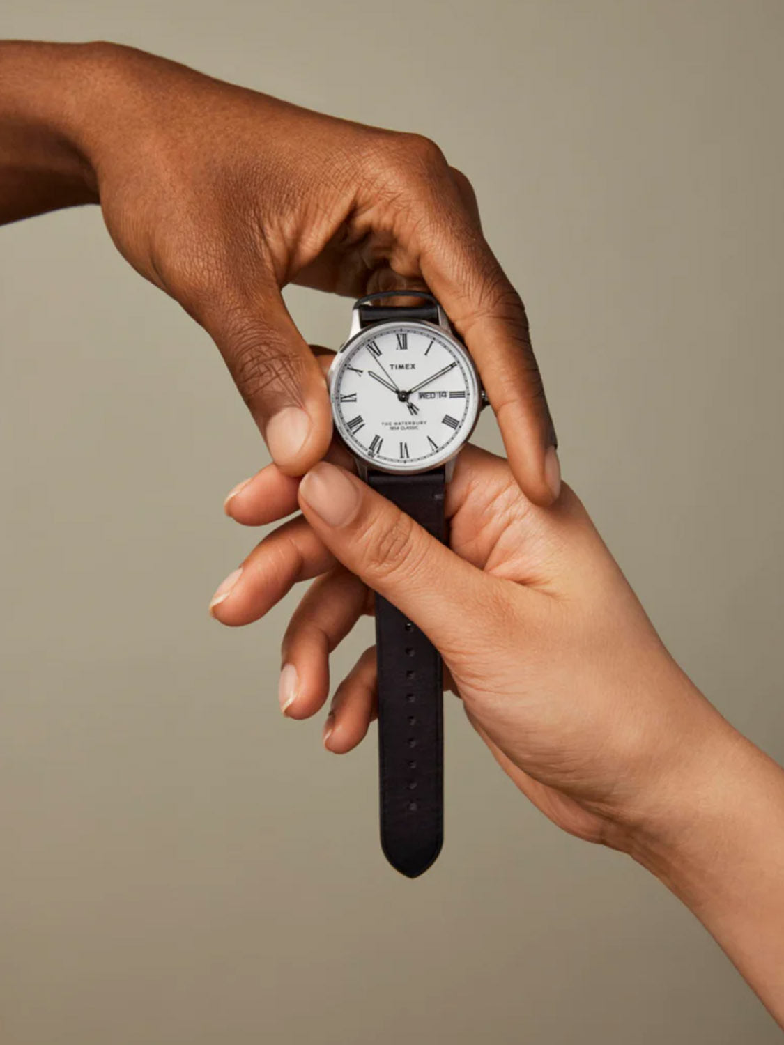 Two hands hold a Timex Waterbury Classic watch with a white face and black quick release leather strap; one hand adjusts the time while the other steadies the watch against a neutral background.