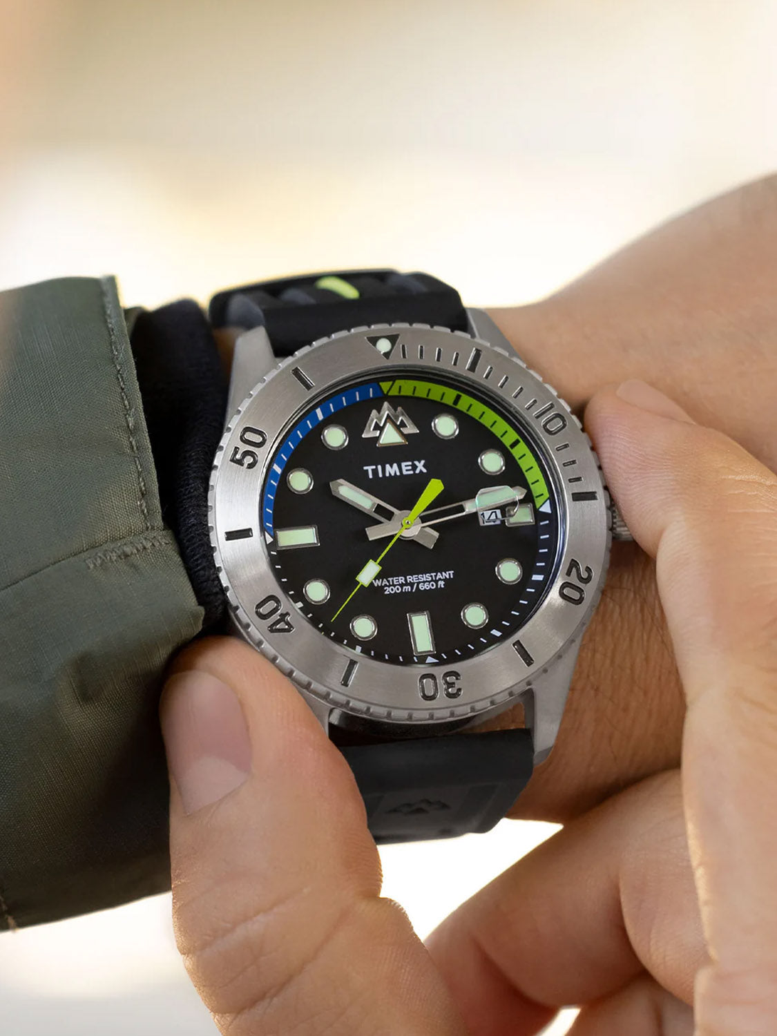 Close-up of someone adjusting a Timex Expedition Anchorage 42mm Silicone Strap Watch. It has a black dial with luminous markers, stainless-steel case, rotatable bezel with minute markings and a green and blue accent ring under the scratch-resistant sapphire crystal, while wearing a green jacket.