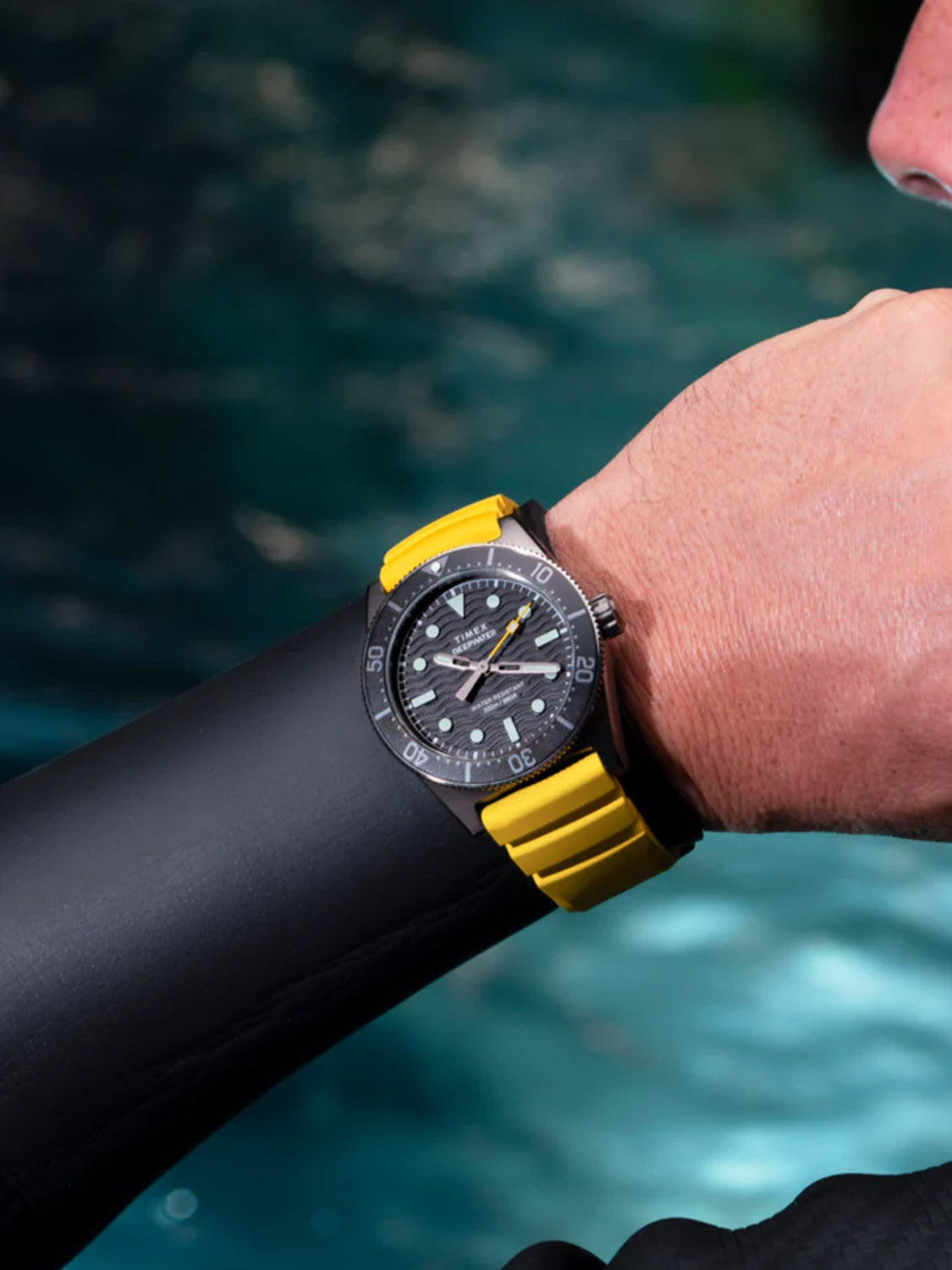 A close-up of a wrist wearing a Timex Deepwater Reef 200 watch, ideal for diving with water resistance. Paired with a sleek black wetsuit, it features Super-Luminova® details against shimmering blue water.