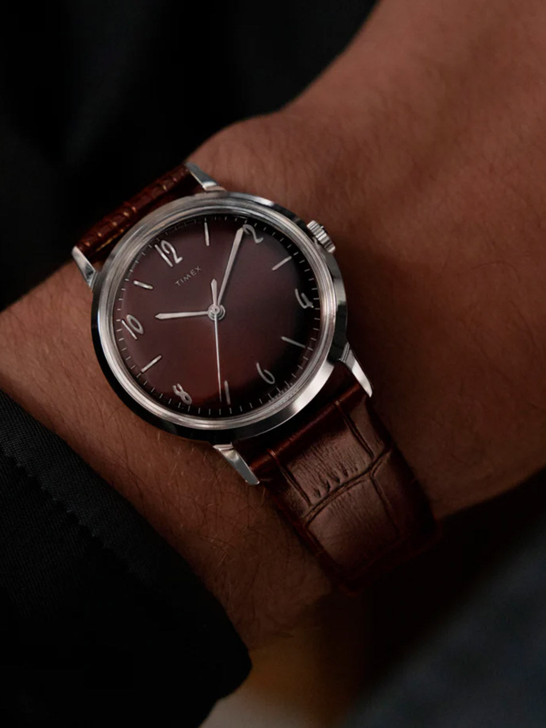 A close-up of a wrist wearing the Timex Marlin® Hand-Wound 34mm Leather Strap Watch, featuring a chocolate brown dial with silver hour markers and hands, paired with an eco-friendly leather strap. The person wears a dark sleeve.