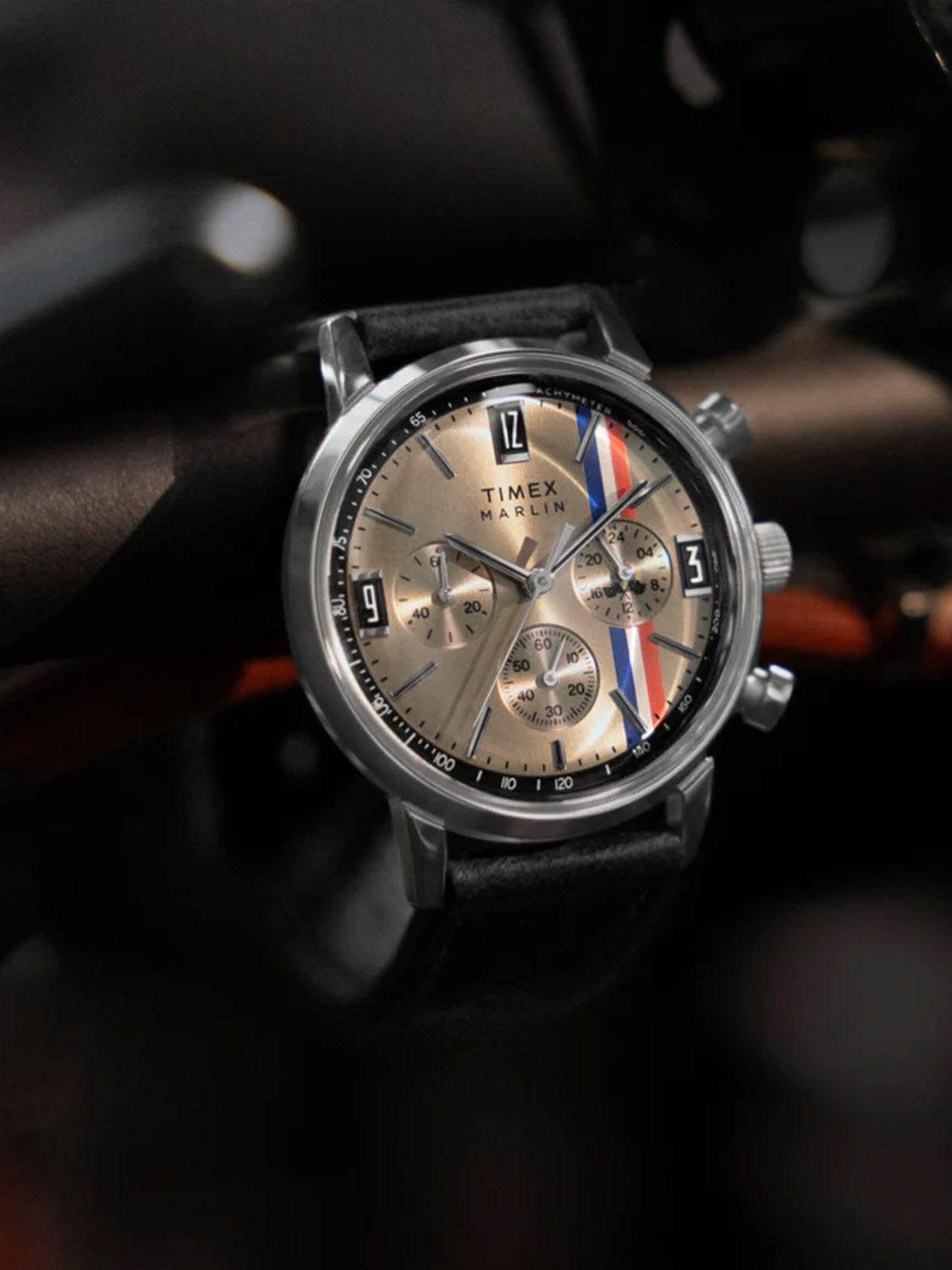 The Timex Marlin® Chronograph Tachymeter features a tan dial with three subdials, a vertical red, white, and blue racing stripe, a black leather strap, and is set against a dark, blurred background.