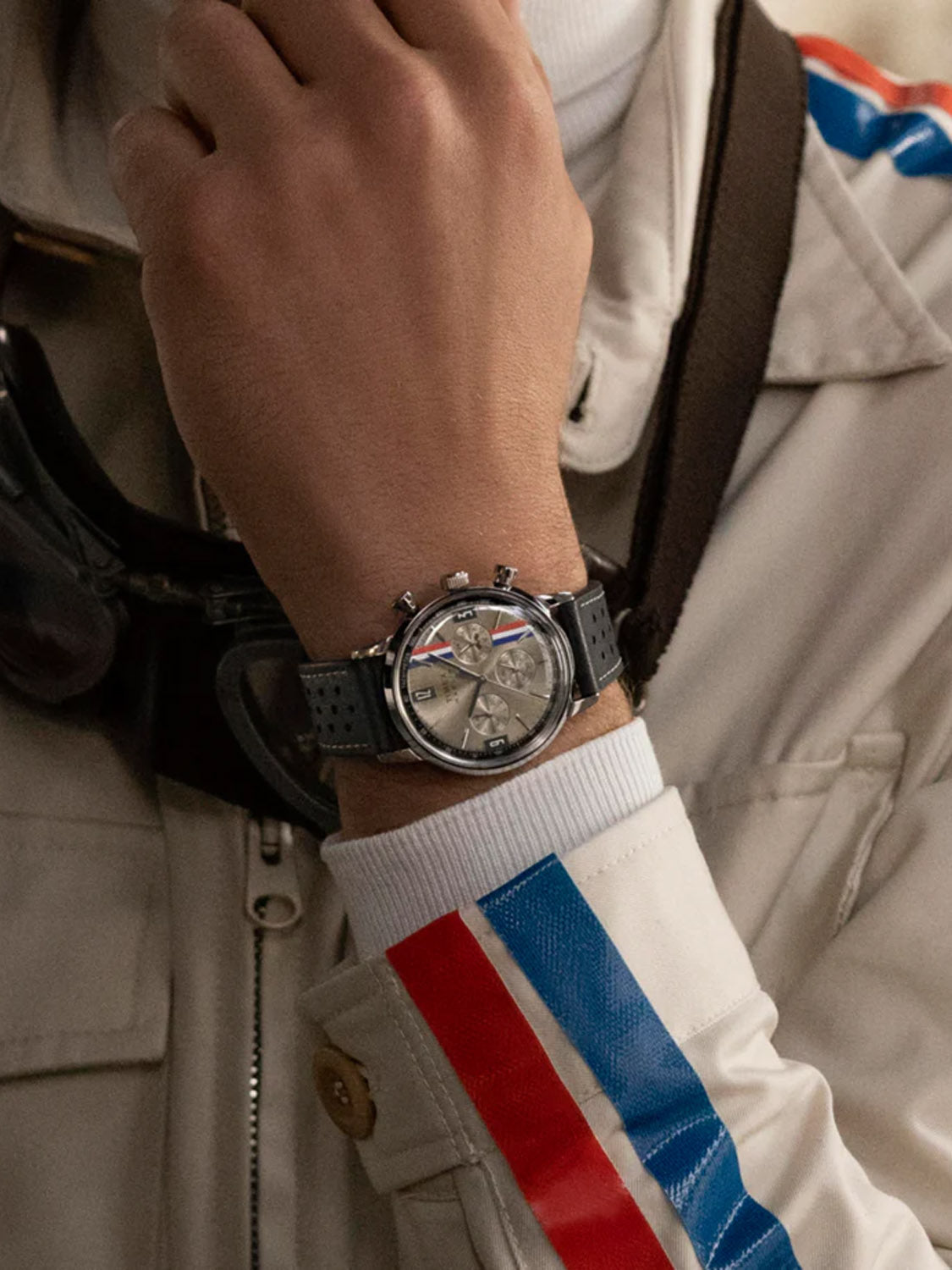 A person in a white jacket with red and blue stripes rests their hand near their chin, displaying a Timex Marlin® Chronograph Tachymeter with a black strap. Racing goggles hang around their neck.