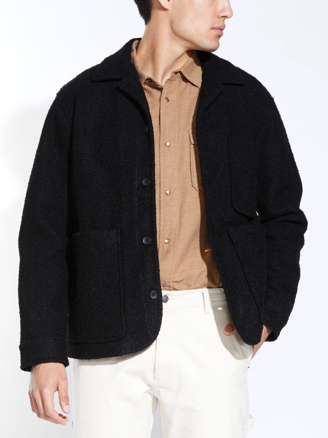 A man models the Civil Society Abbott Relaxed Fit Walking Coat in black, layered over a tan button-down shirt and white pants, standing with one hand in his pocket against a plain white background.