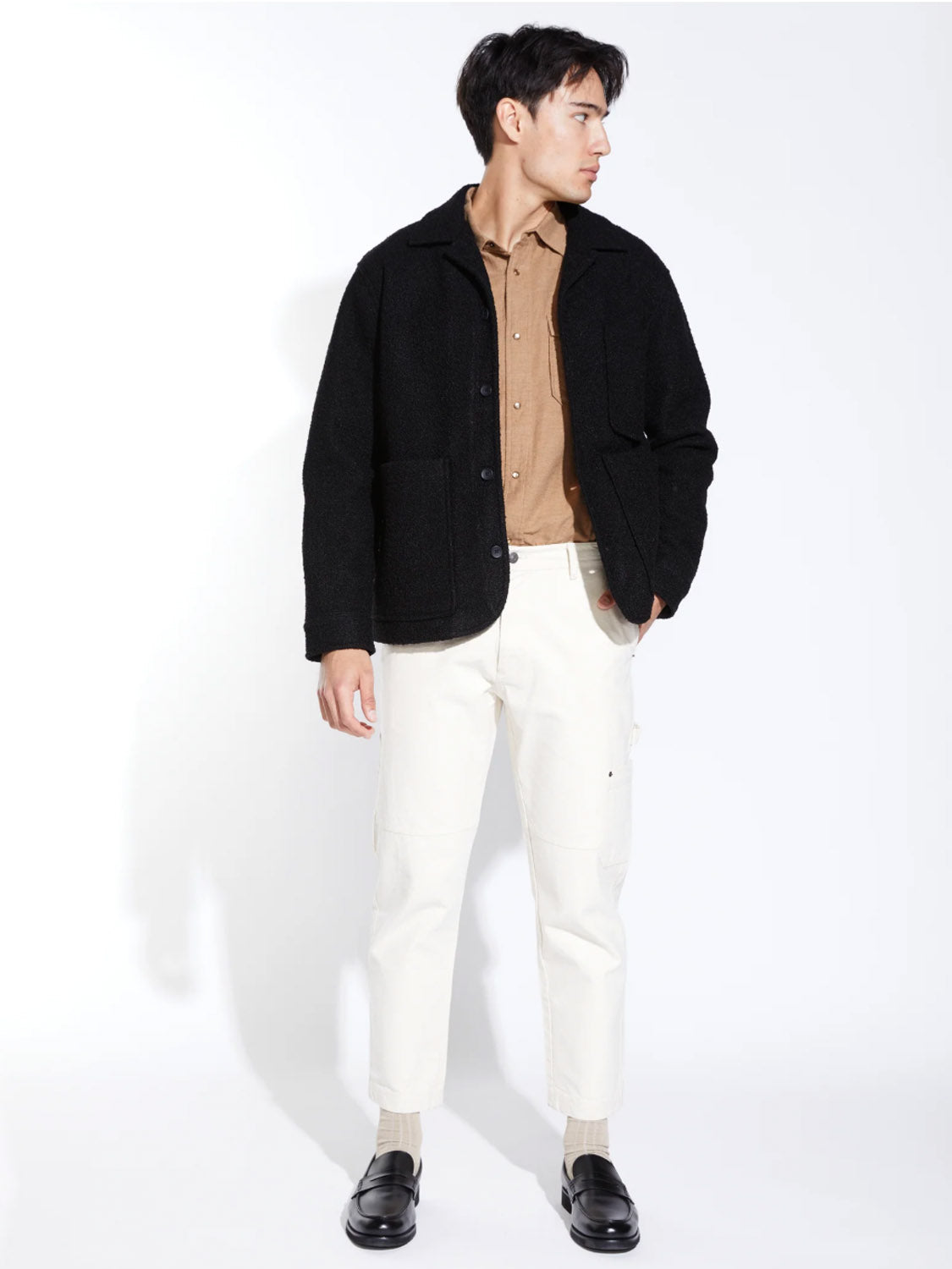 A man stands against a white background, wearing the Civil Society Abbott Relaxed Fit Walking Coat in black over a tan button-up shirt, paired with white pants and black loafers. He looks to his left with one hand in his pocket.