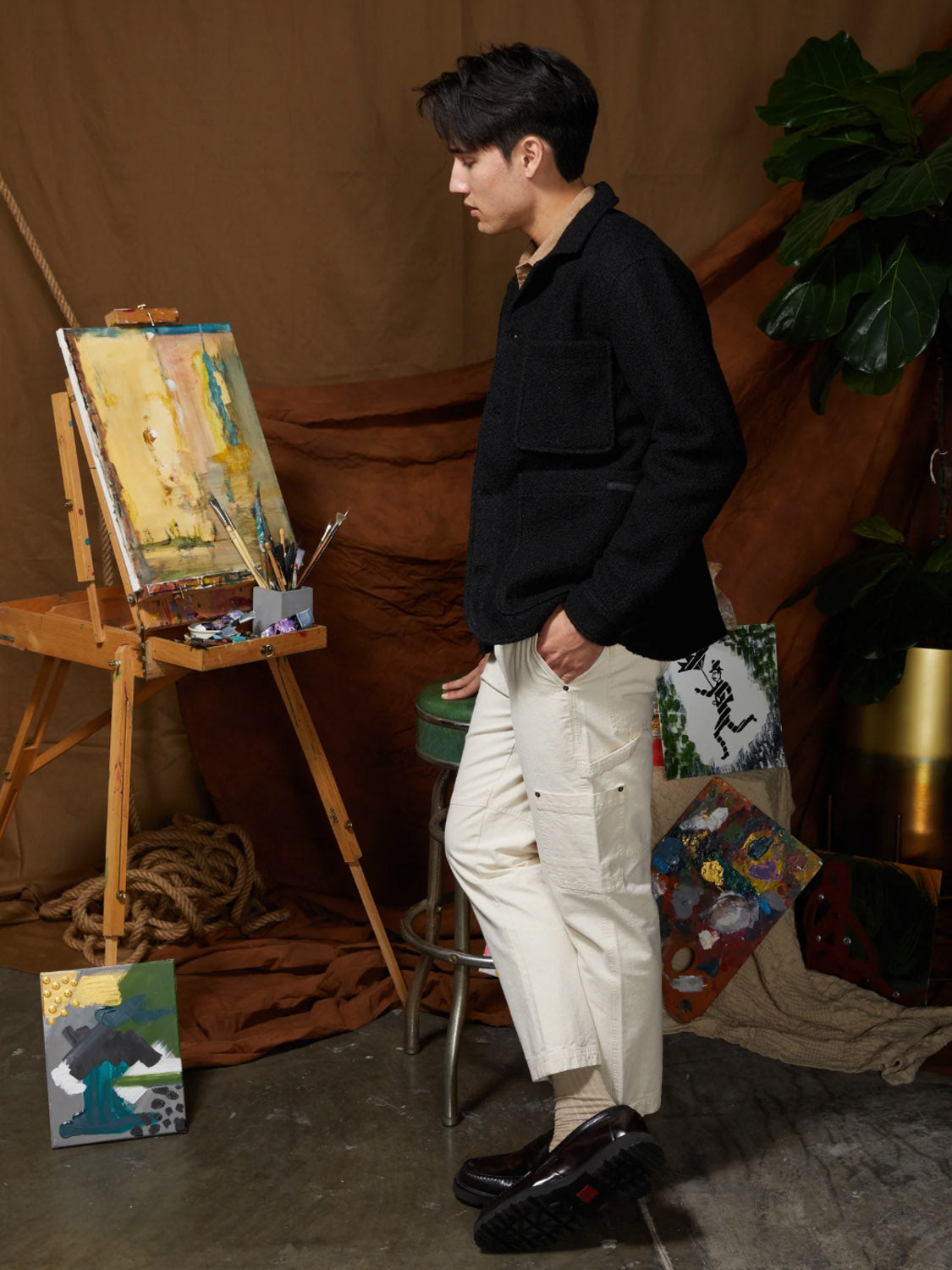 A person wearing the Civil Society Abbott Relaxed Fit Walking Coat in black and white pants stands beside an easel with a colorful painting in a studio, surrounded by art supplies, finished paintings, and large green leaves.