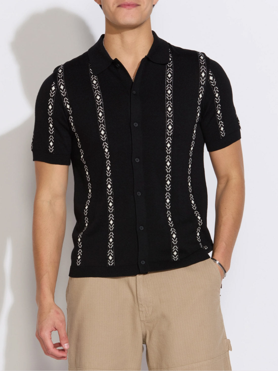 A man wears the Ace Full Placket Sweater Polo by Civil Society—a retro black short-sleeve button-up knit shirt with vertical white jacquard stripes—paired with light beige pants, standing against a plain white background.