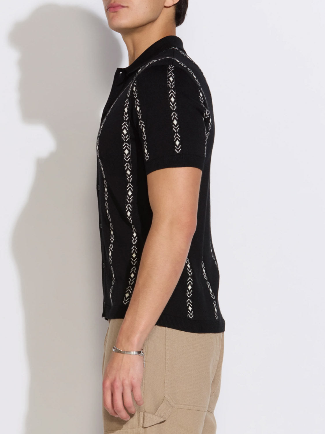 A person stands in profile wearing the Civil Society Ace Full Placket Sweater Polo, a retro short-sleeved black knit shirt with vertical white jacquard stripes, paired with light brown pants against a plain light background.