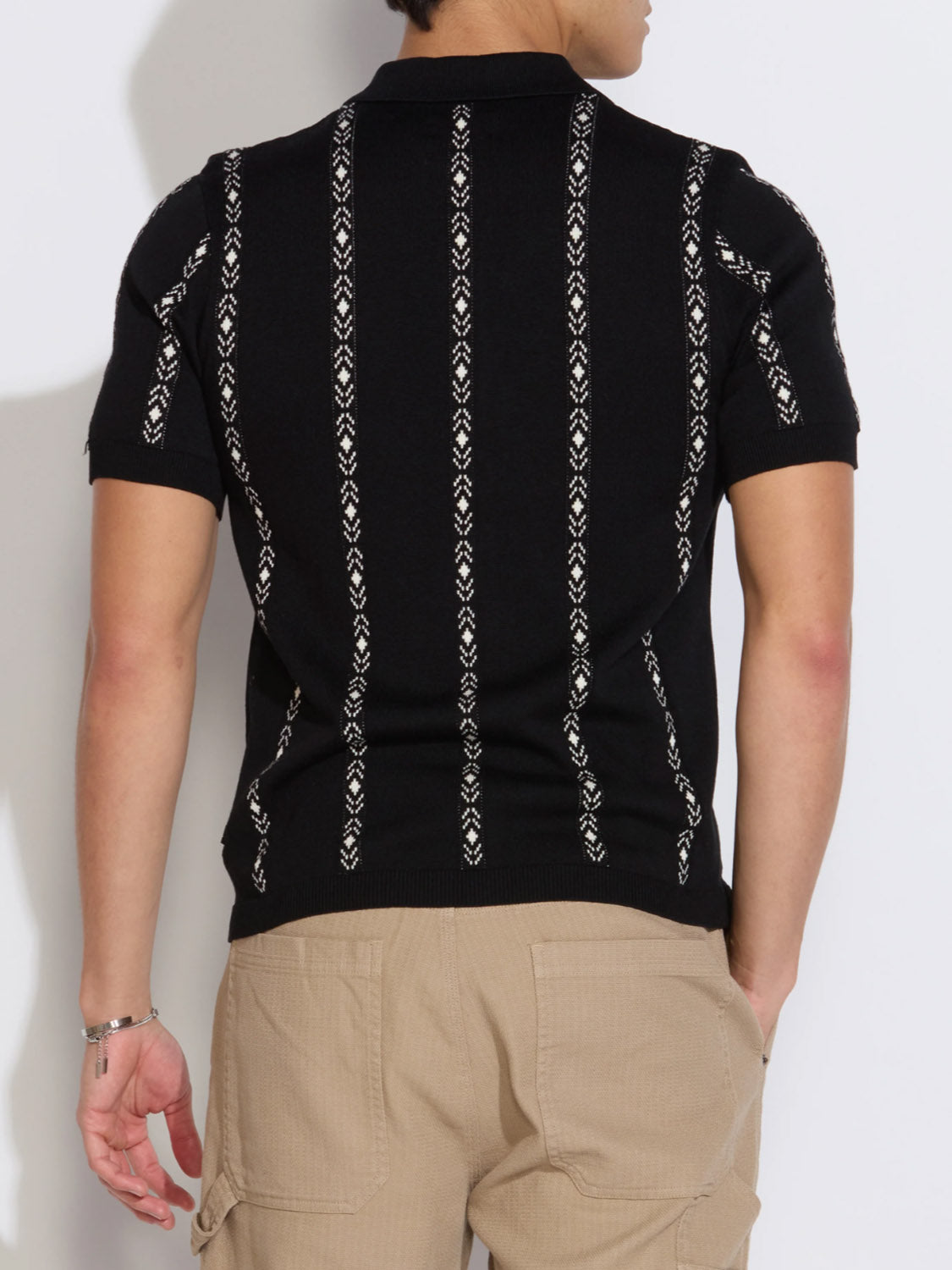 A person with short dark hair, seen from the back, wears the Civil Society Ace Full Placket Sweater Polo—a black retro short-sleeve shirt with white jacquard stripes—paired with beige cargo pants and a silver bracelet on their left wrist.