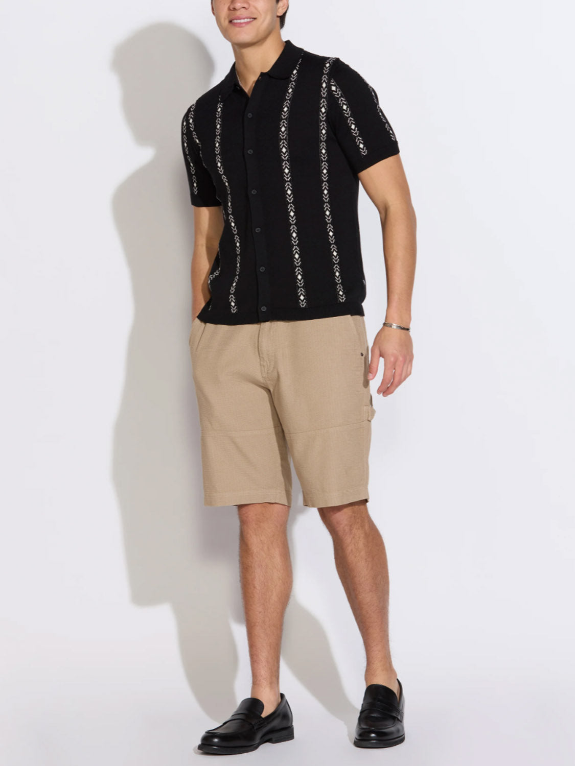 A man stands against a white background wearing the Civil Society Ace Full Placket Sweater Polo, styled with tan knee-length shorts and black loafers for a touch of retro flair.
