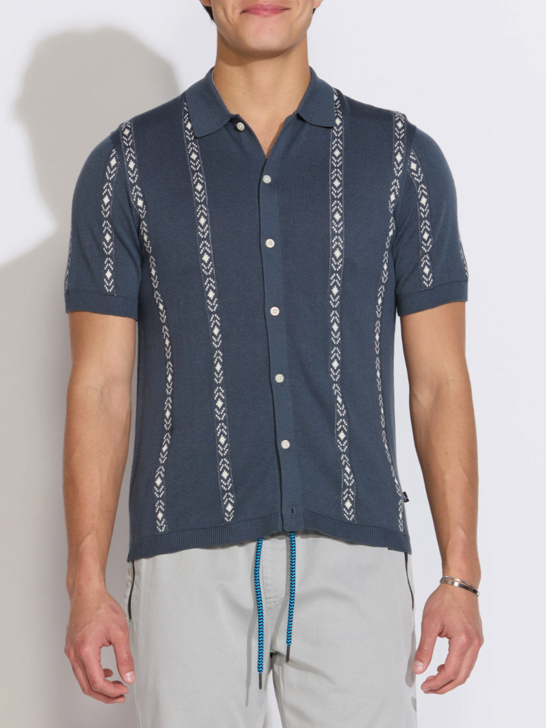 A man wears the Civil Society Ace Full Placket Sweater Polo, a short-sleeve dark blue jacquard stripe shirt, paired with light gray pants with a blue drawstring, standing against a plain white background.