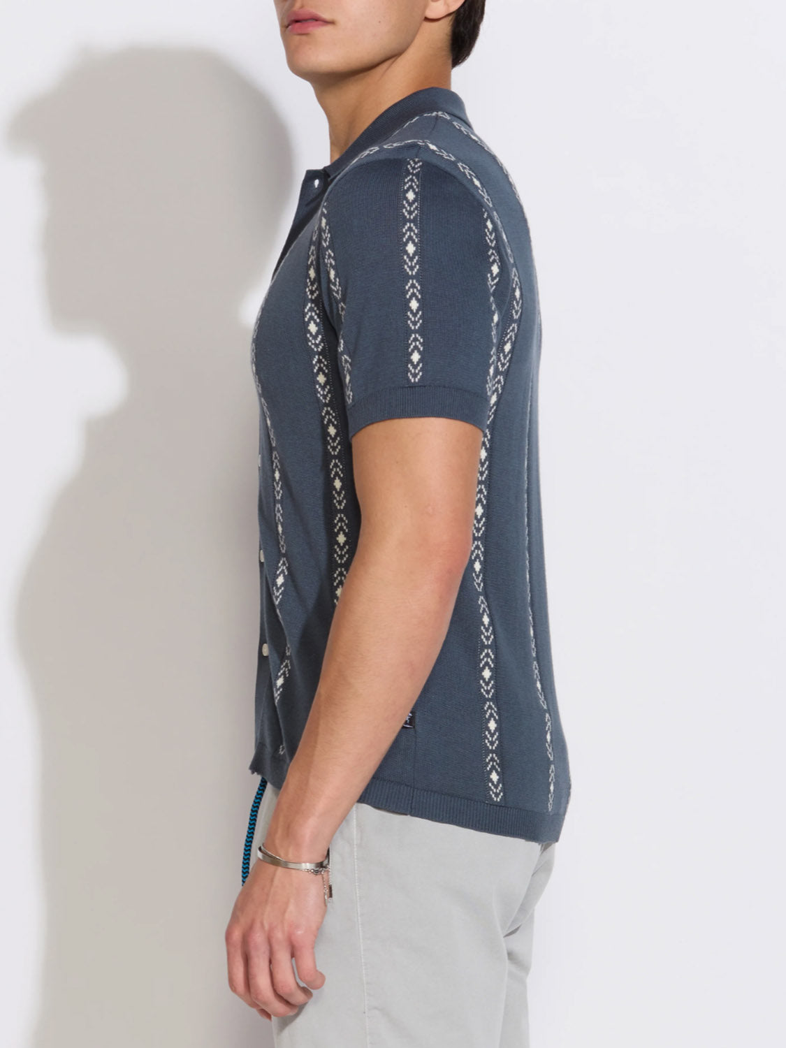 A person in profile wears the Civil Society Ace Full Placket Sweater Polo, featuring blue jacquard stripes, paired with light gray pants for a subtle retro look against a plain white background.