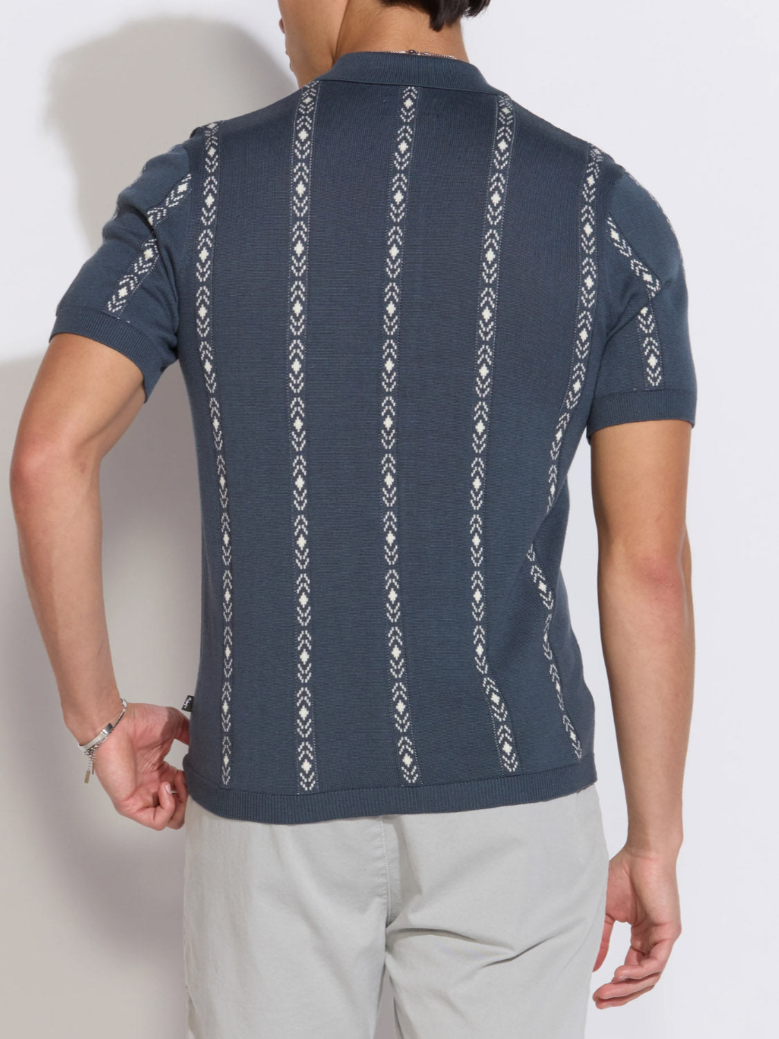 A person is seen from the back wearing the Civil Society Ace Full Placket Sweater Polo in dark blue with vertical jacquard stripes and light gray pants. The short sleeves and bent right arm stand out against a plain background.