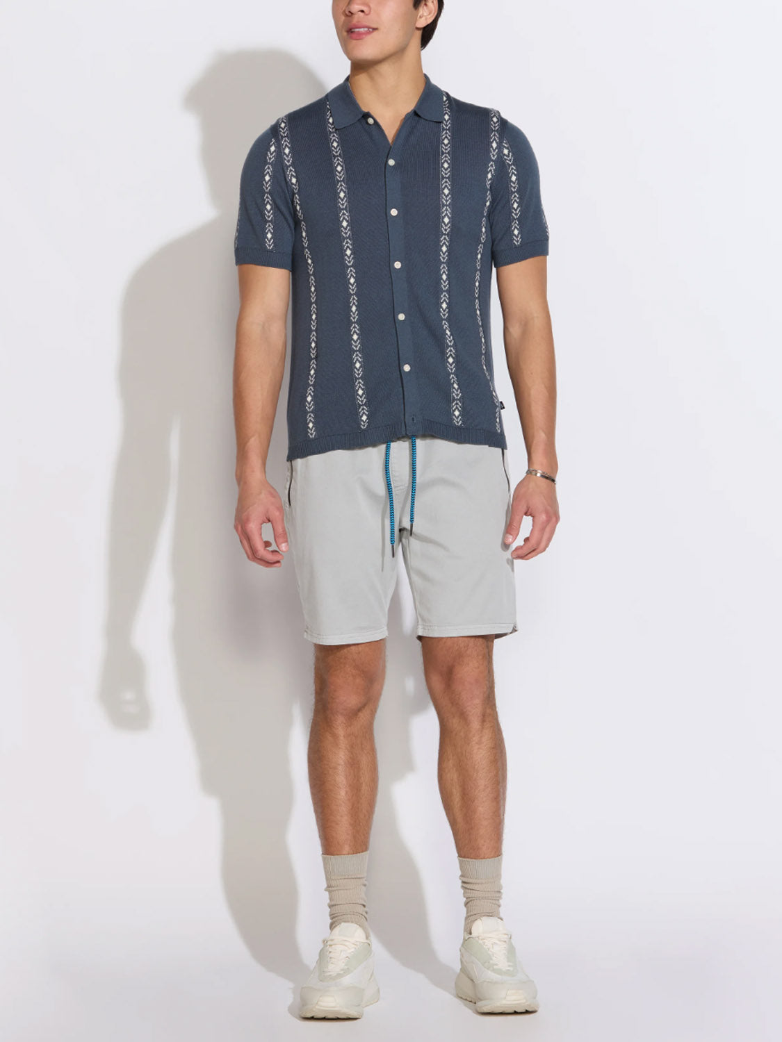 A man faces forward against a white background, wearing the Civil Society Ace Full Placket Sweater Polo in navy with light gray shorts, blue drawstring, beige socks, and white sneakers for a stylish retro look.