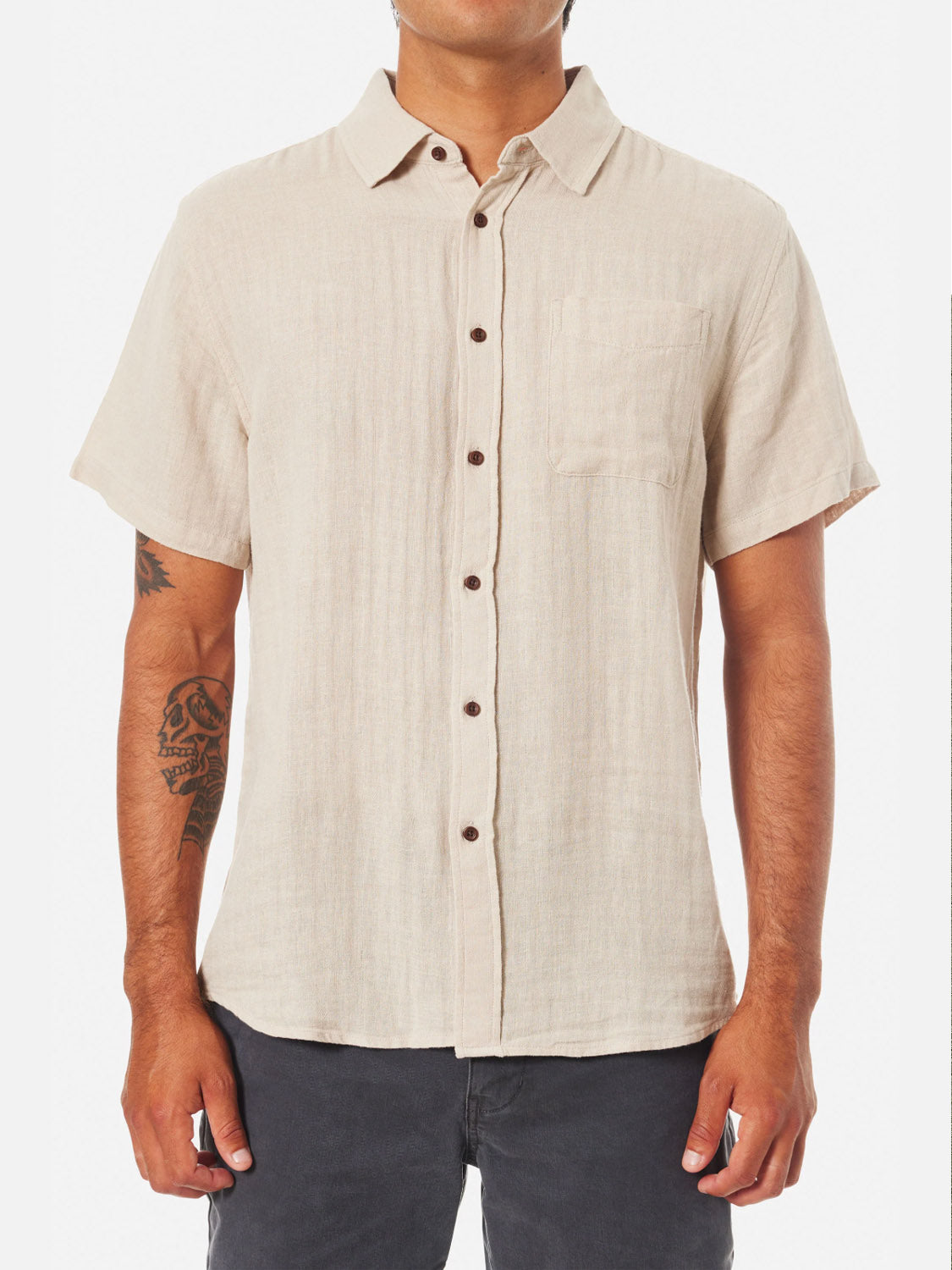 A person in the Katin Alan Solid Shirt by Katin—a short-sleeve beige button-up—stands facing forward. Sporting a visible right forearm tattoo and dark pants, they highlight versatile style against a plain white background.