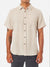A person in the Katin Alan Solid Shirt by Katin—a short-sleeve beige button-up—stands facing forward. Sporting a visible right forearm tattoo and dark pants, they highlight versatile style against a plain white background.