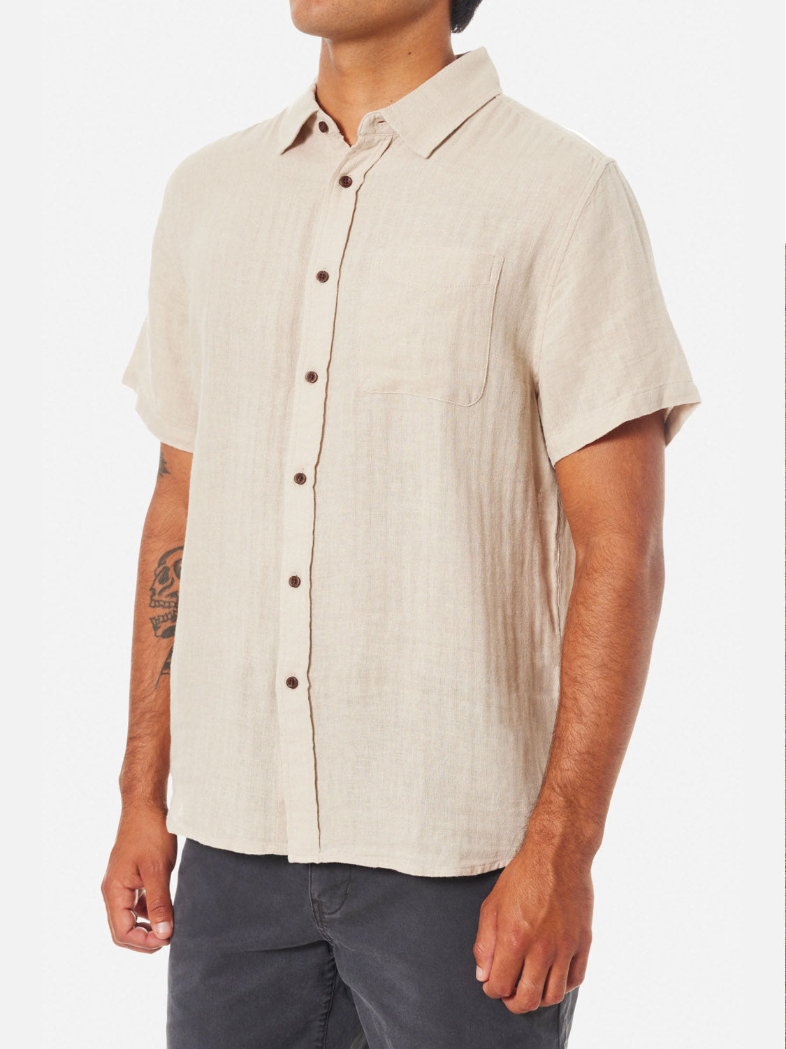 A man wears the Katin Alan Solid Shirt by Katin, a beige short-sleeve button-up with a chest pocket and dark buttons. He matches it with dark pants, revealing a left forearm tattoo, against a plain white background.